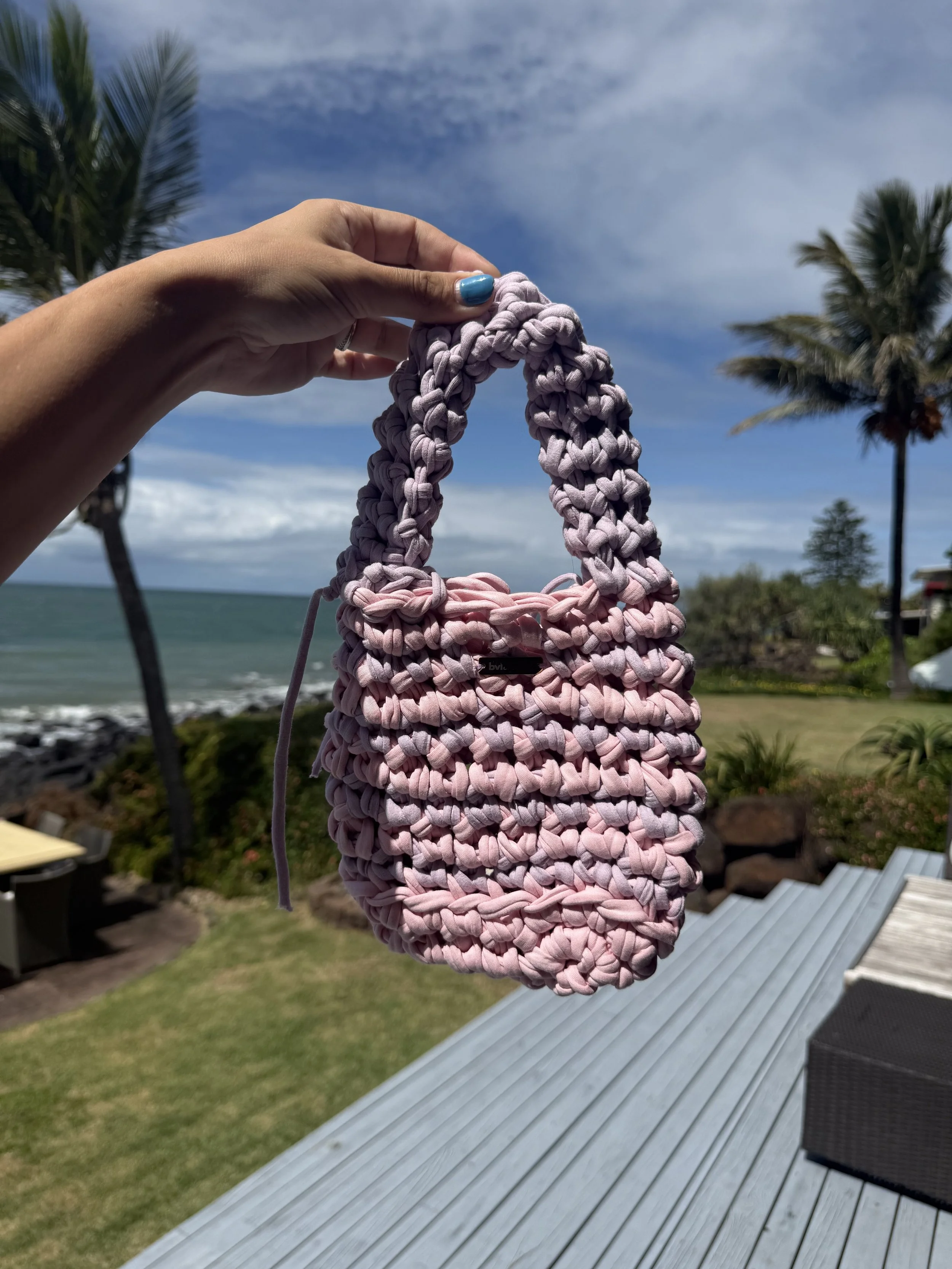 Close-up of a hand with blue painted nails holding a pink woven bag outdoors by the beach, with palm trees, grassy area, ocean, and partly cloudy sky in the background.