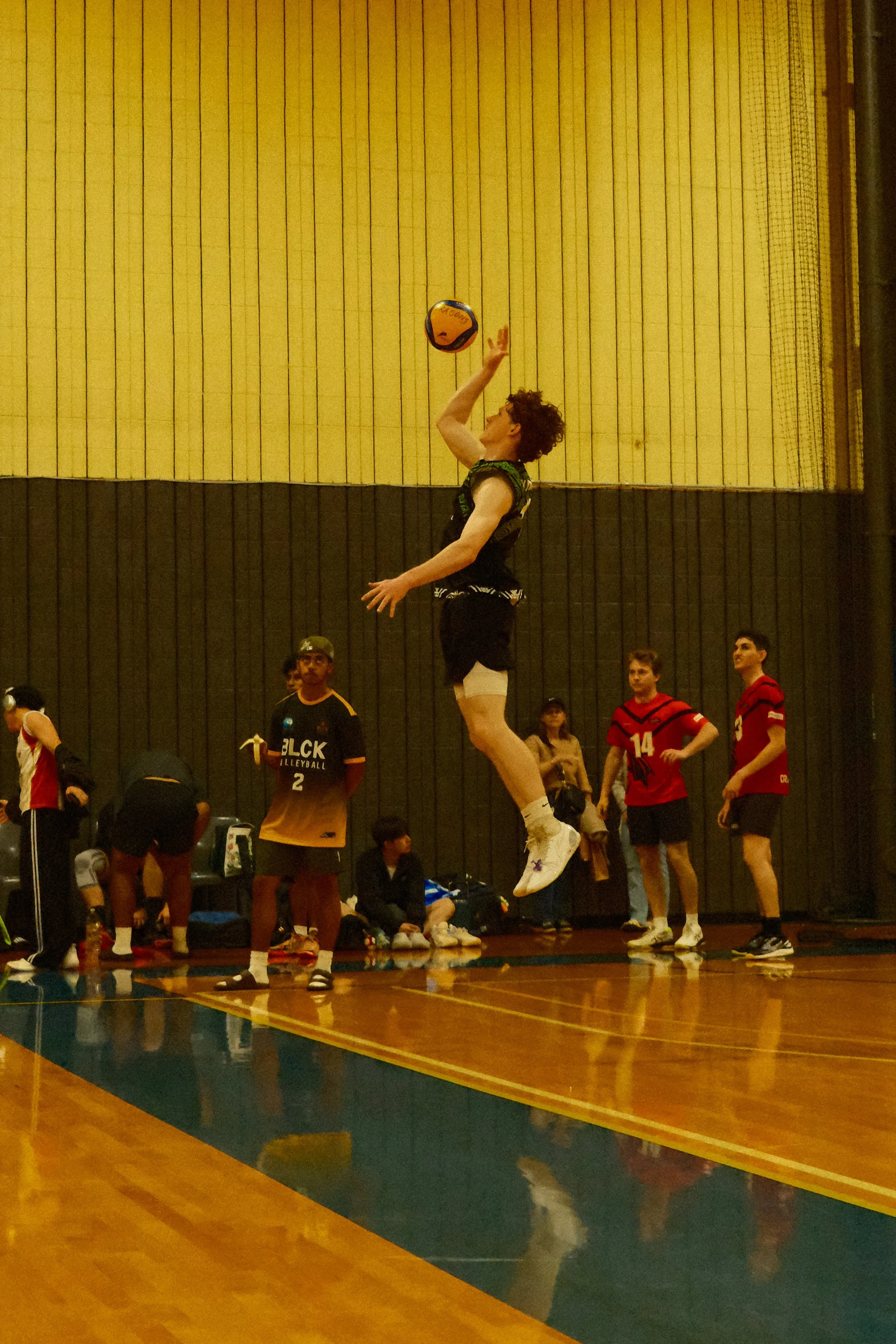 A person is jumping in a volleyball game, reaching to hit the ball, with several spectators watching in an indoor gymnasium with yellow and dark-colored walls.