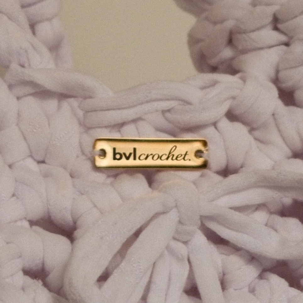 Close-up of a white, chunky crocheted item with a gold tag that reads 'bvl crochet' attached to it.