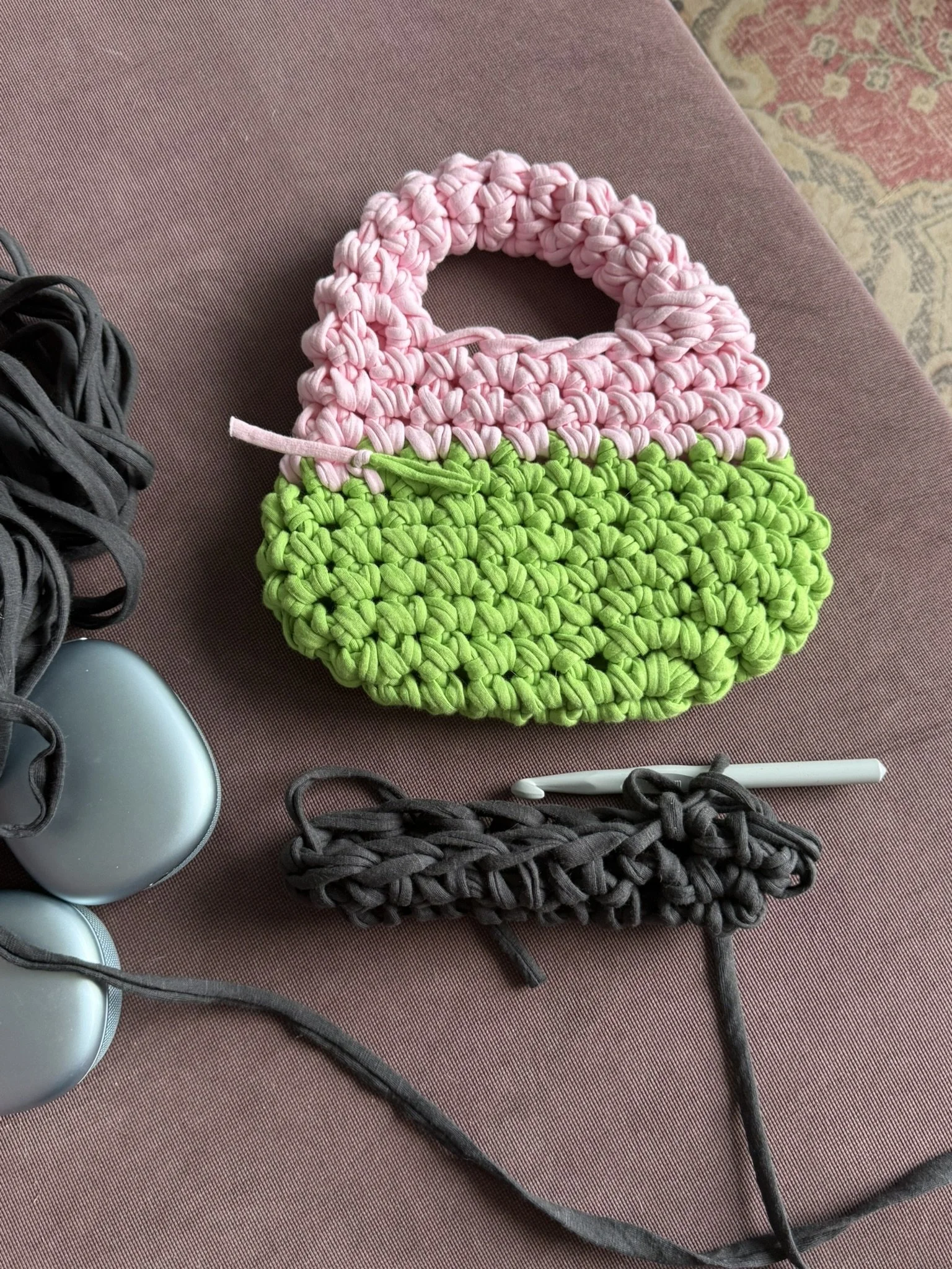 Three handmade woven bags in pink, green, and dark gray on a brown surface, with gray shoes and a pen nearby.