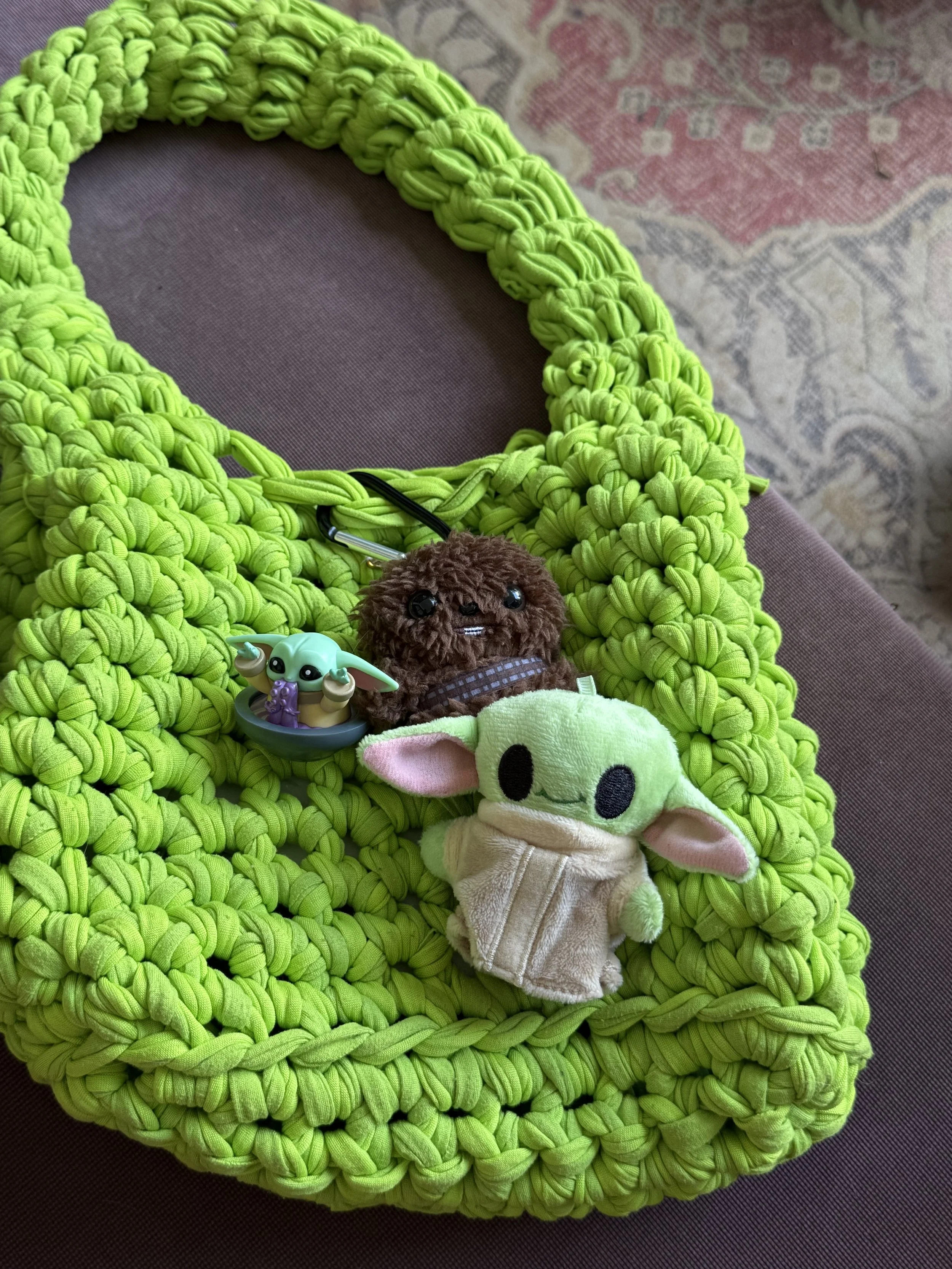 Bright green hand-knit blanket with plush toys on top, including a Baby Yoda plush, a brown teddy bear, and a small plush with large ears and black eyes, on a dark brown surface near a patterned rug.