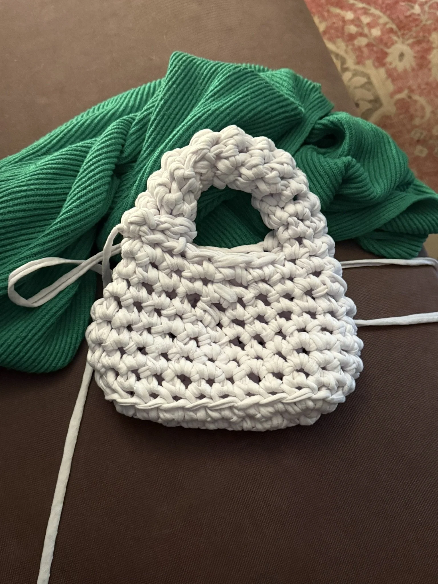 A white crocheted bag rests on a brown surface, with a green corduroy fabric and a portion of an ornate patterned rug visible in the background.
