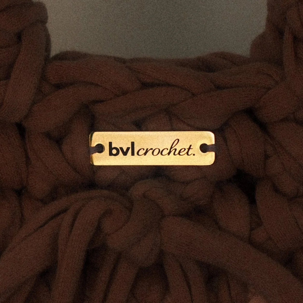Close-up of a gold-colored tag with the text 'bvlcrochet.' attached to a brown, braided, knitted fabric.