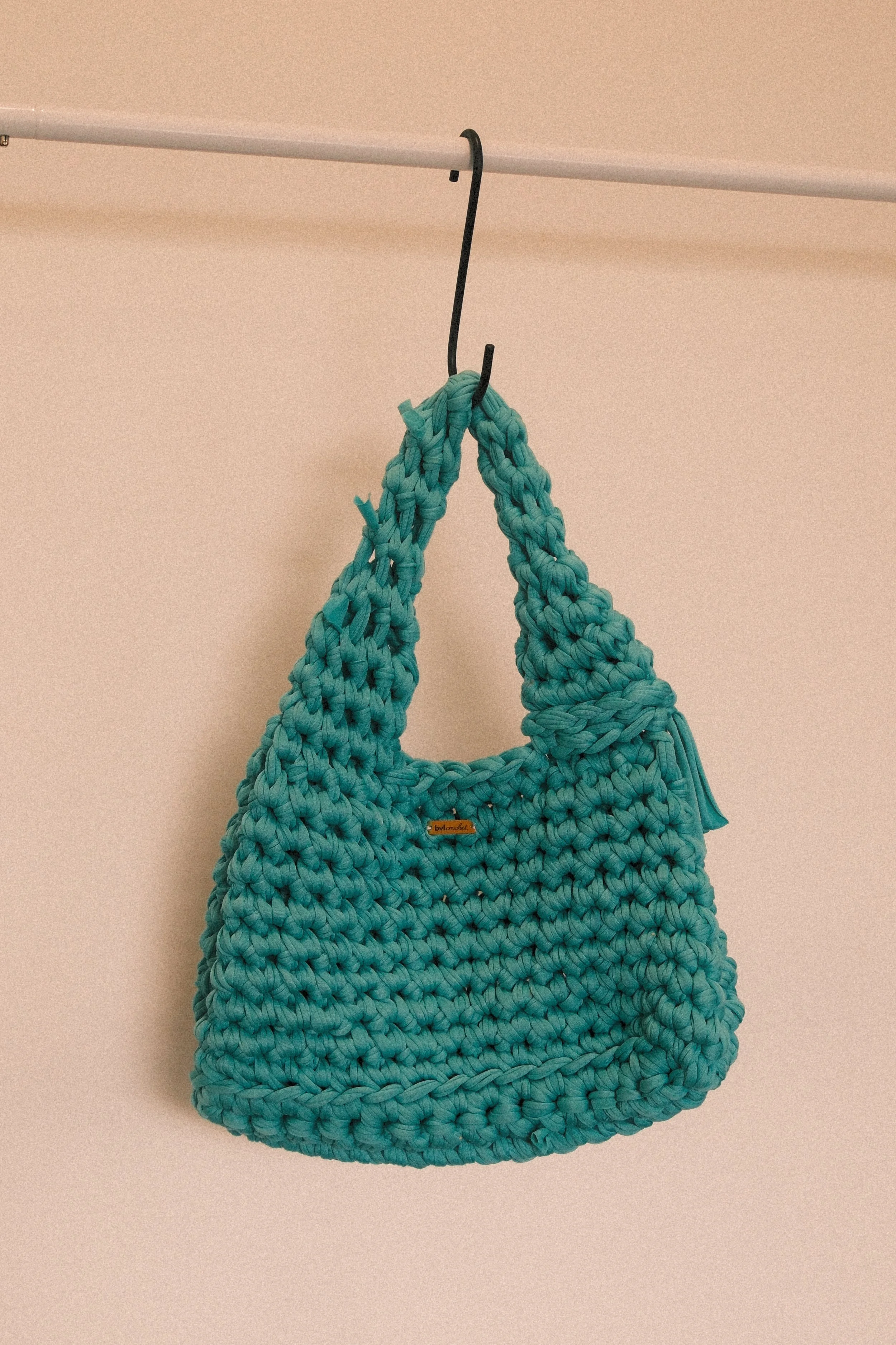A turquoise crocheted handbag hanging from a black hook on a beige wall.