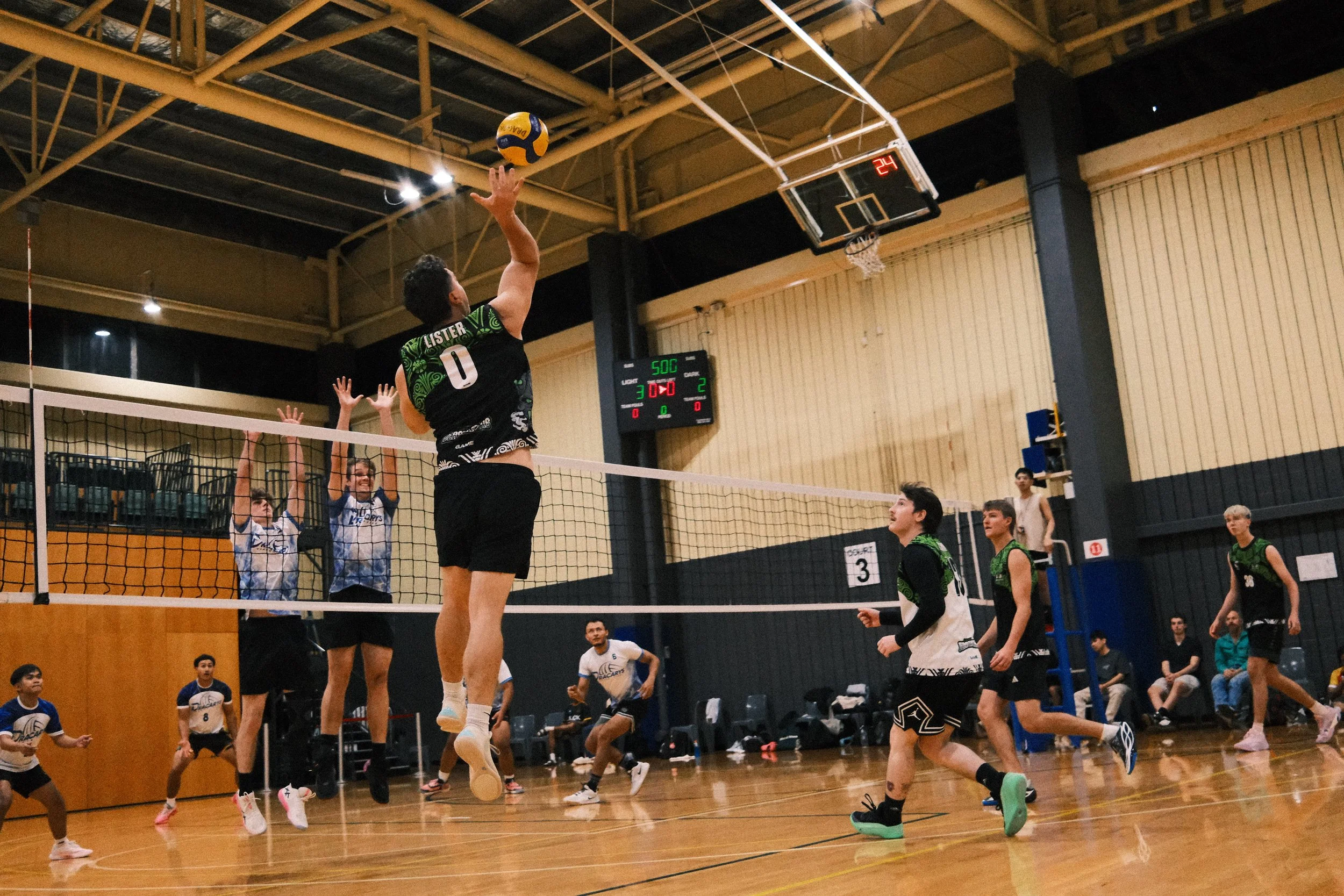 A volleyball player in a black and green jersey jumps to hit the ball over the net during a game in an indoor gymnasium. Multiple players on both teams are visible, with some preparing to react to the play. The gym has a wooden floor and a scoreboard displaying the score and time.
