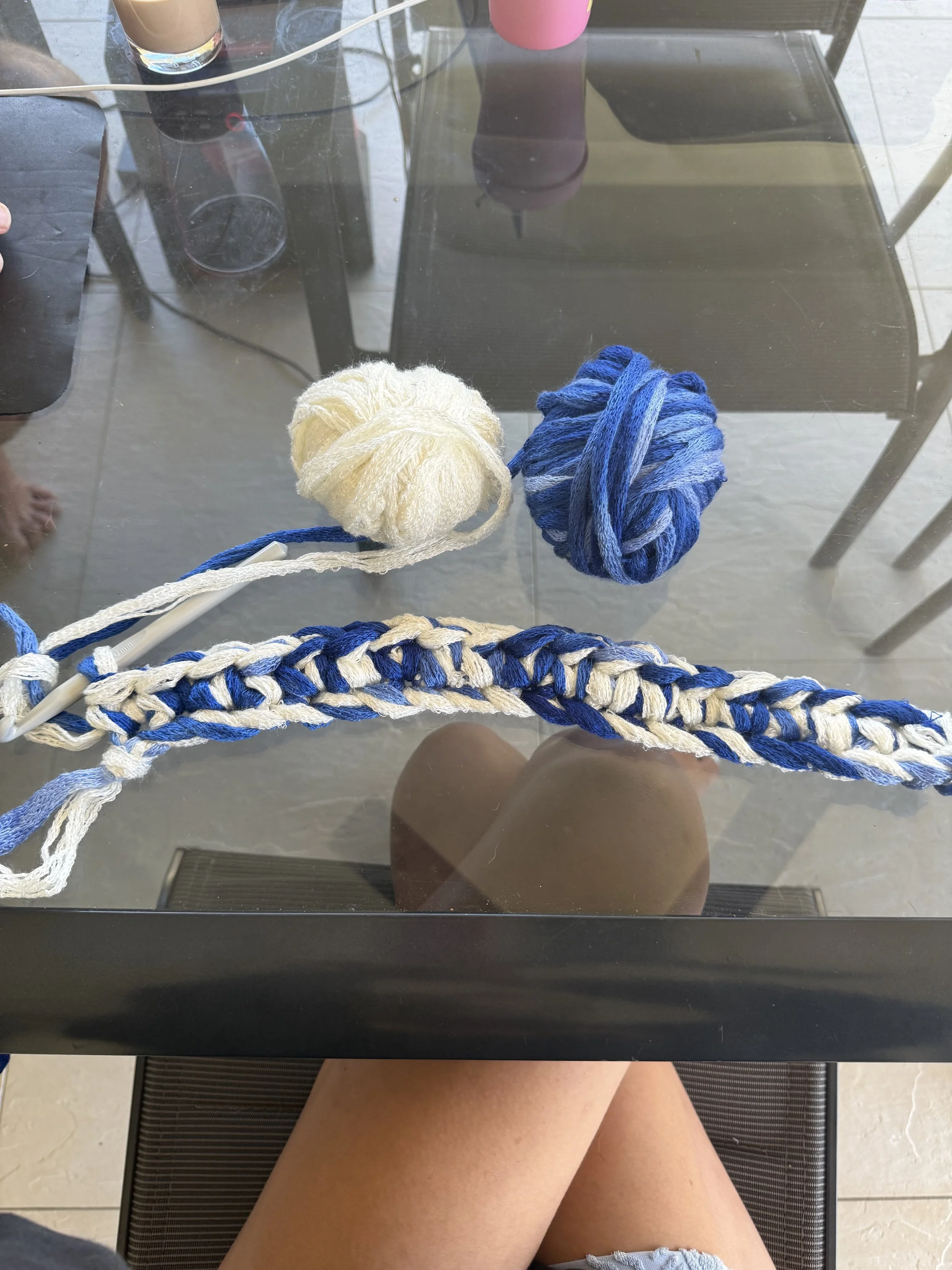 Top-down view of a glass table with two yarn balls, one cream and one blue, resting on a crochet project. A person's legs are visible underneath the table, sitting on a patio with a tiled floor.