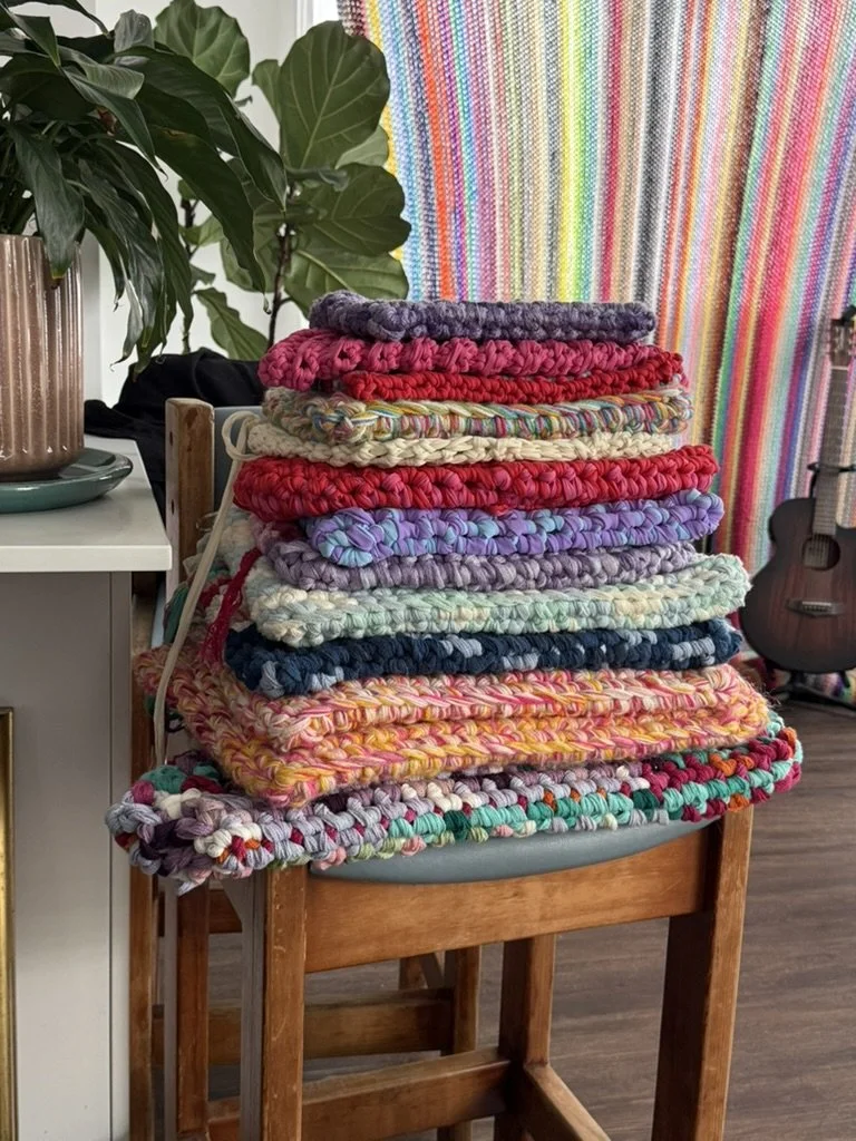 A stack of colorful knitted or crocheted blankets on a wooden chair in a cozy room with a rainbow-striped curtain, a guitar, and a potted plant in the background.