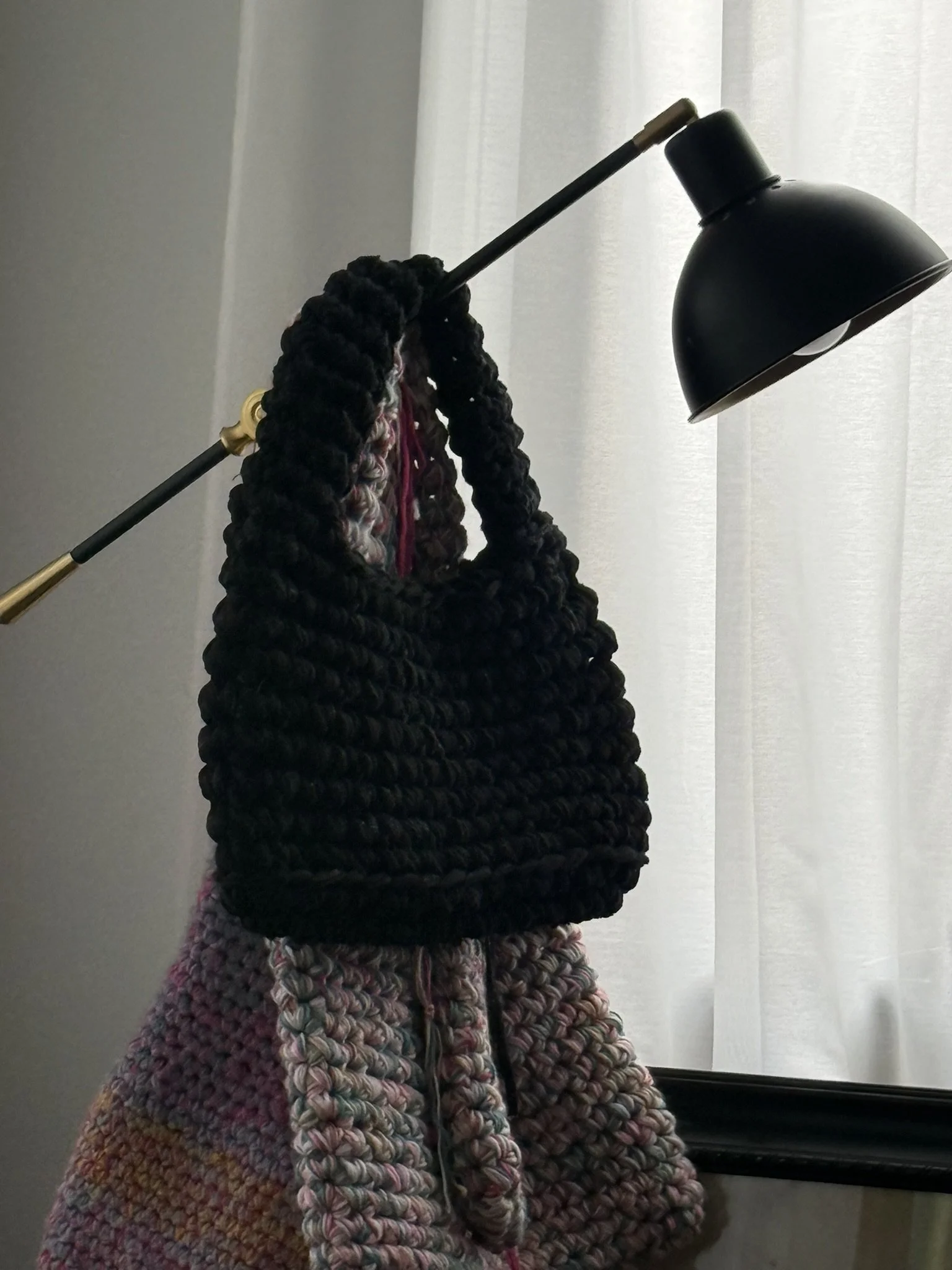 A person wearing a multicolored, chunky, knitted coat and a black knitted hat with a wide brim, holding a black and gold lamp, in front of a white curtain and a window ledge.