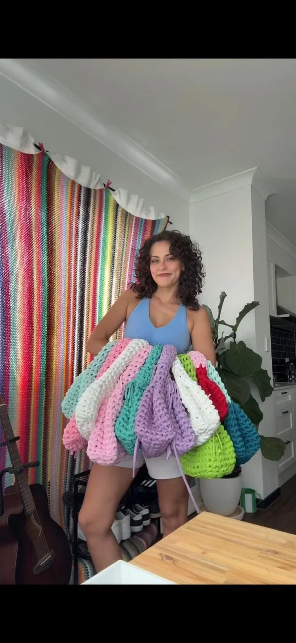A woman with curly dark hair wearing a blue tank top holding multiple colorful crocheted bags in a room with a colorful striped curtain, a guitar, and green plants.