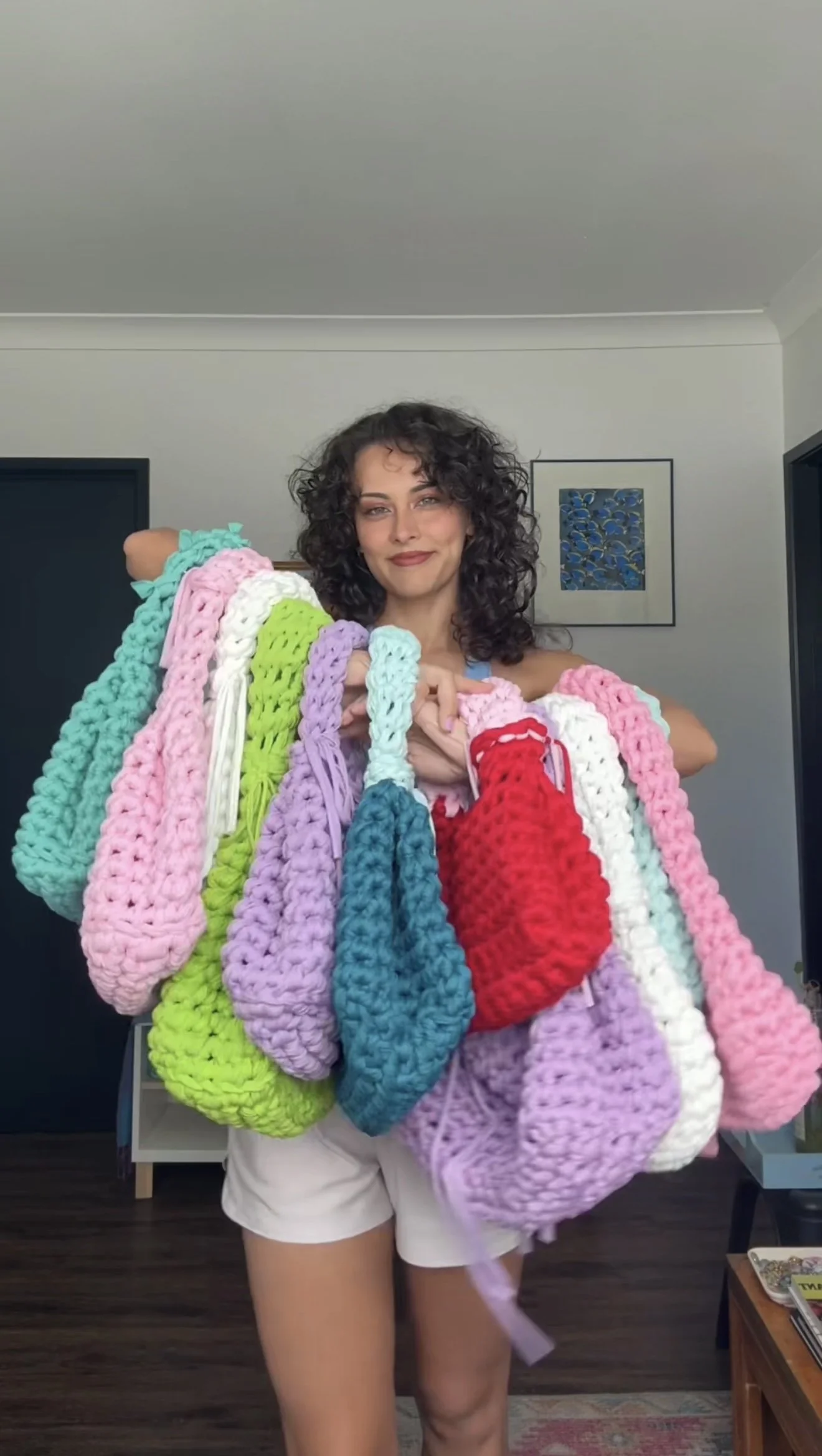 Woman with curly hair smiling, holding multiple colorful chunky knit scarves in a room with artwork on the wall.