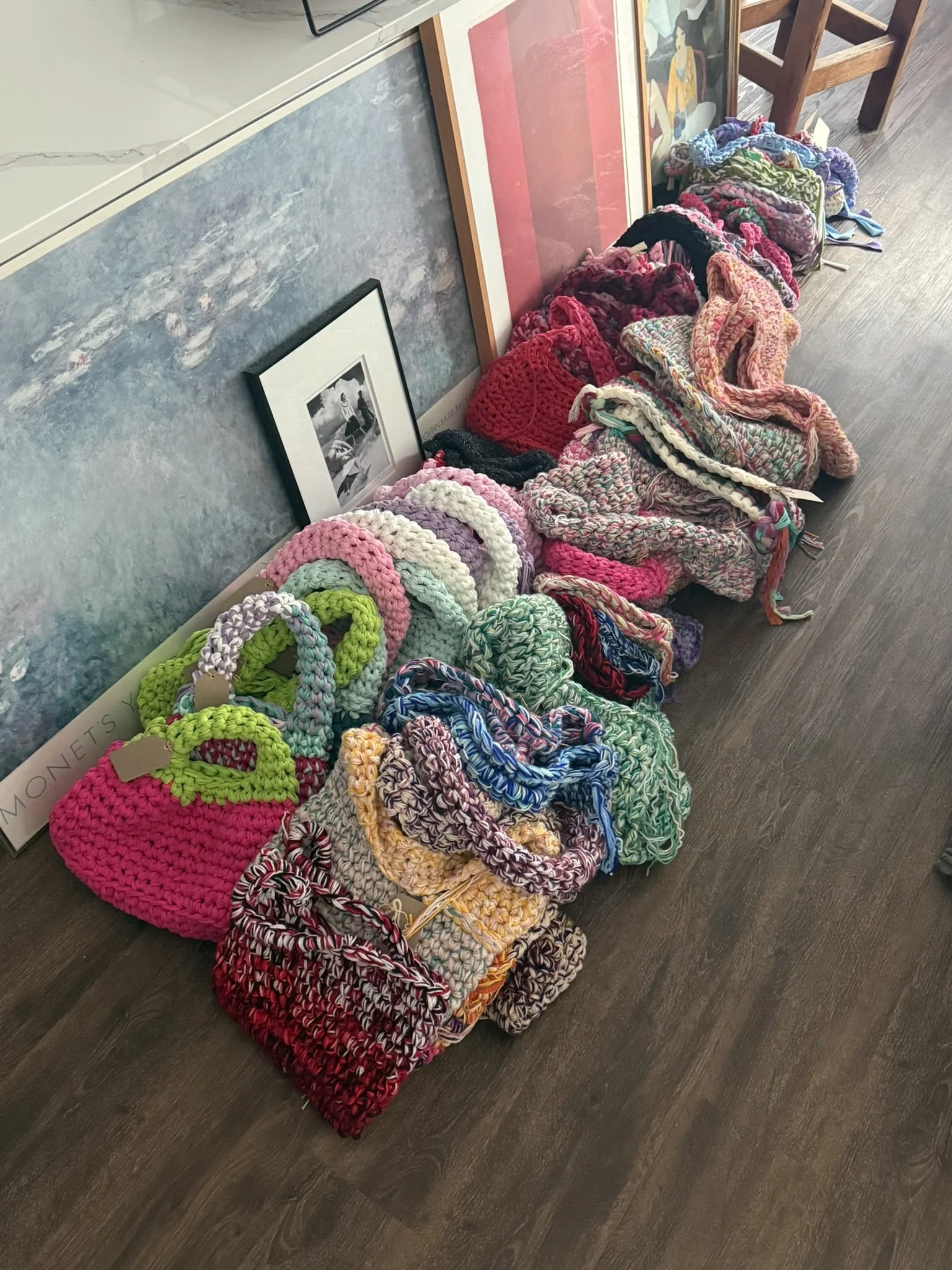 Colorful handmade crocheted bags arranged on the floor next to framed artwork and a wooden chair, with additional framed artwork leaning against the wall.
