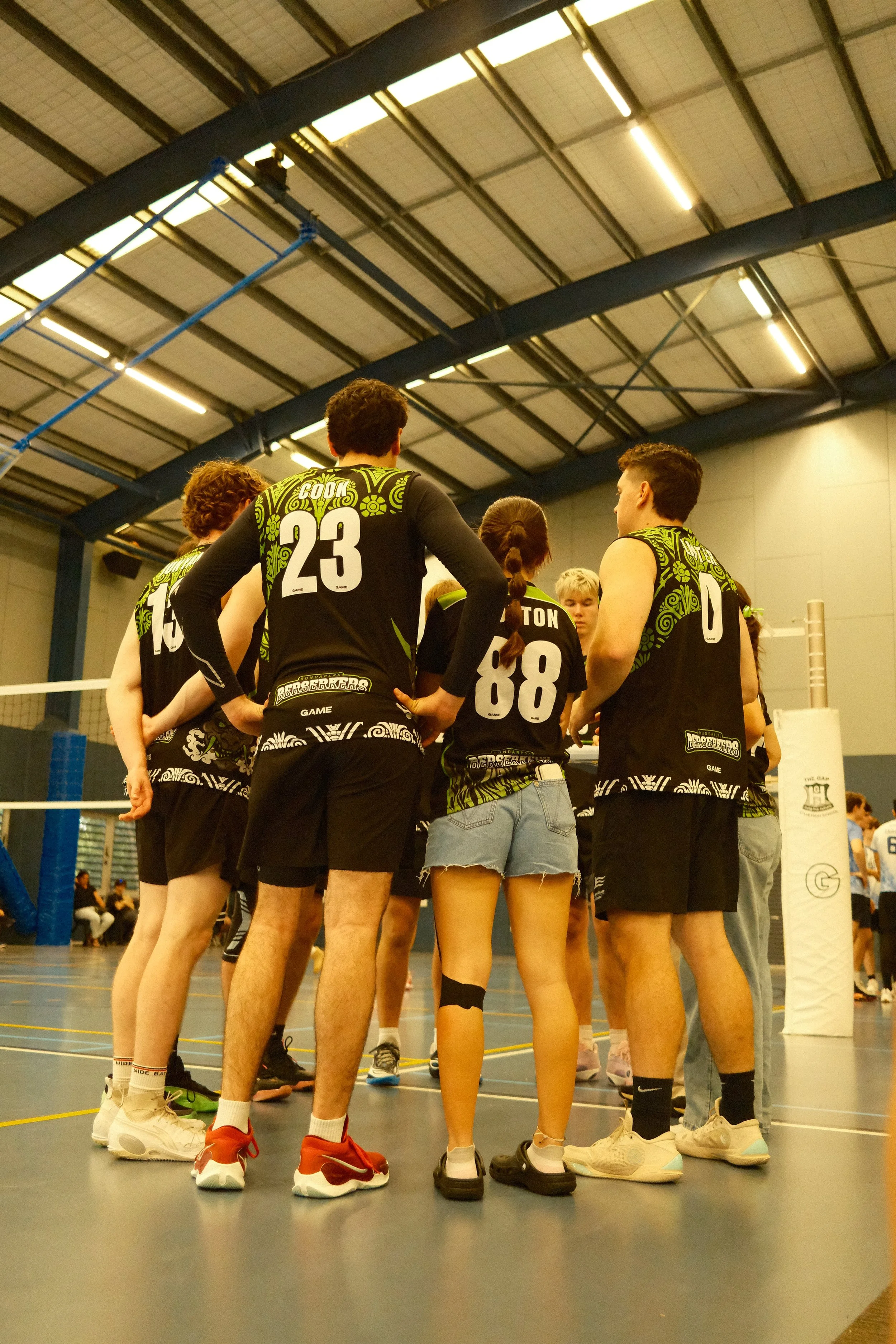 A group of volleyball players huddling together on an indoor court, wearing black and green uniforms with numbers on the back, listening to their coach during a game or practice.