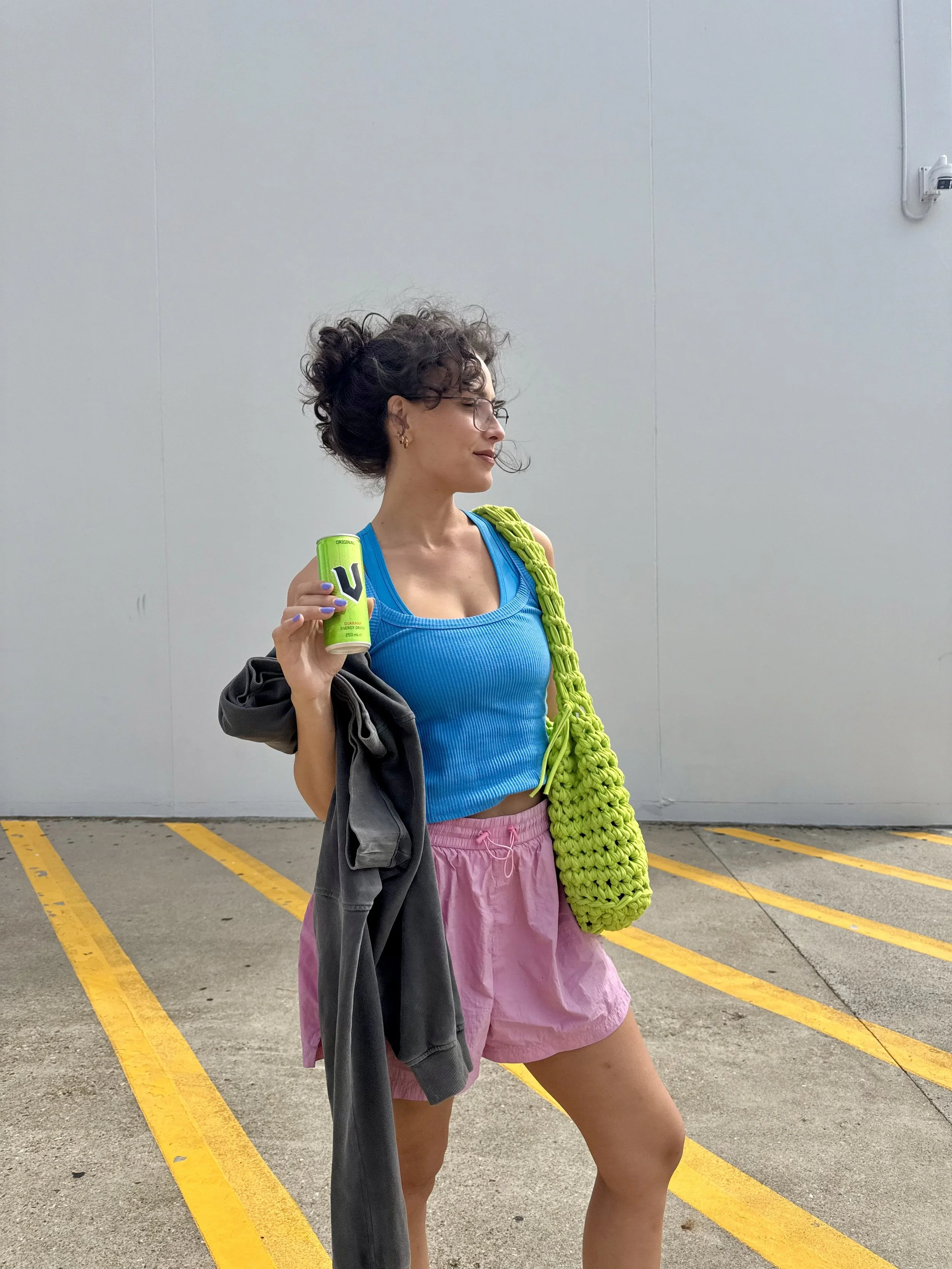 A woman with glasses and curly hair, wearing a blue tank top and pink shorts, holding a can of energy drink, standing in an outdoor parking lot with yellow lines on the ground.
