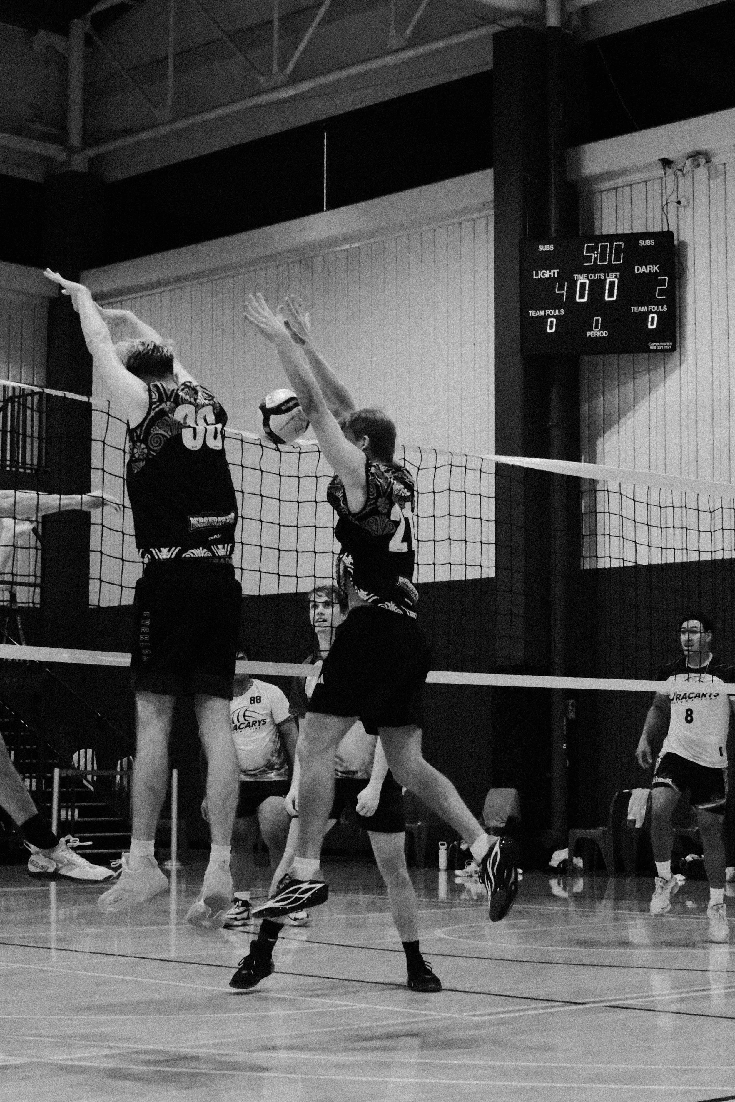 Indoor volleyball game with players jumping at the net. Players are wearing sports jerseys and athletic shoes. Scoreboard shows 5-0 with 40 seconds on the clock.