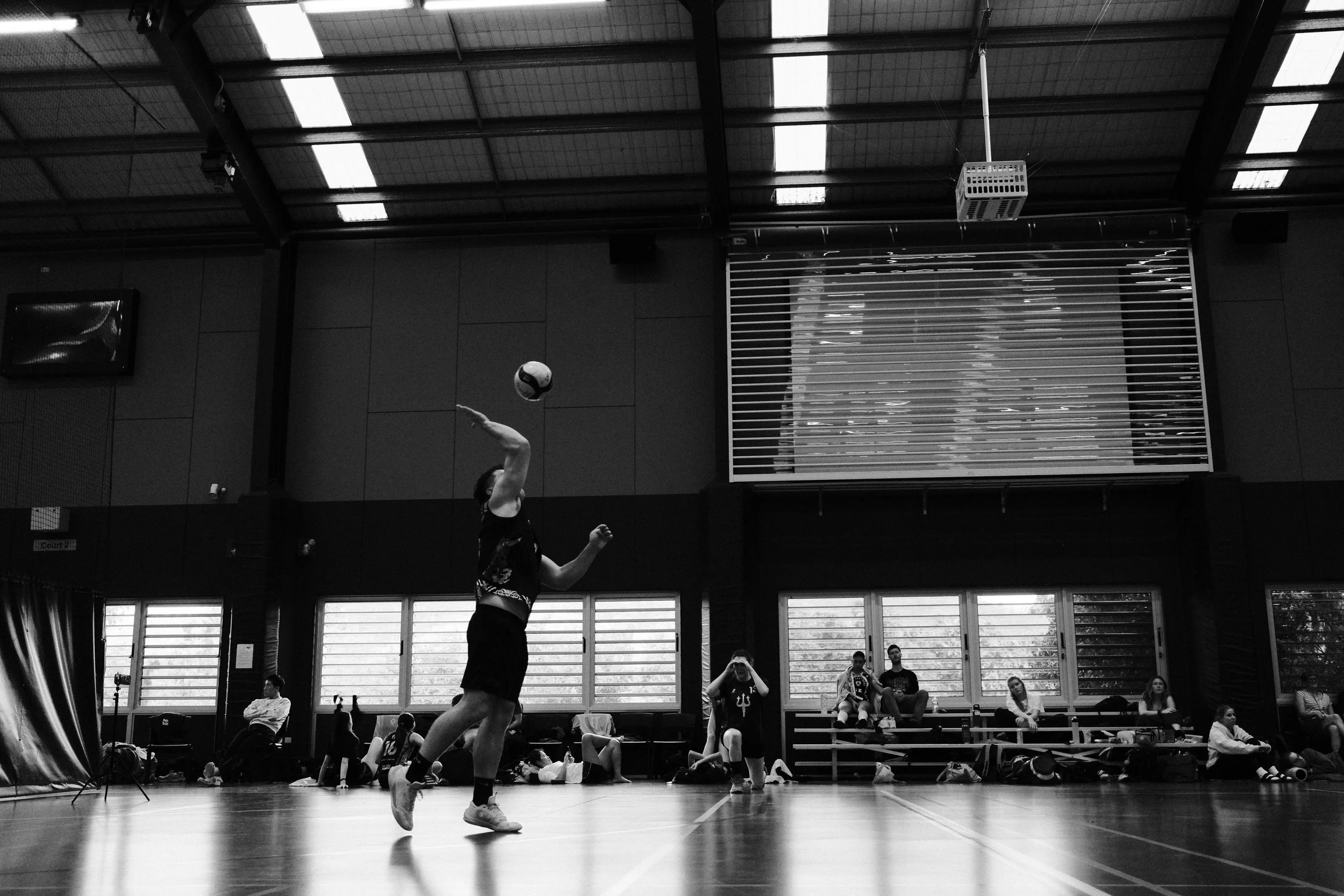 A person playing volleyball inside a gymnasium, jumping to hit the ball, with a group of spectators and players watching in the background.
