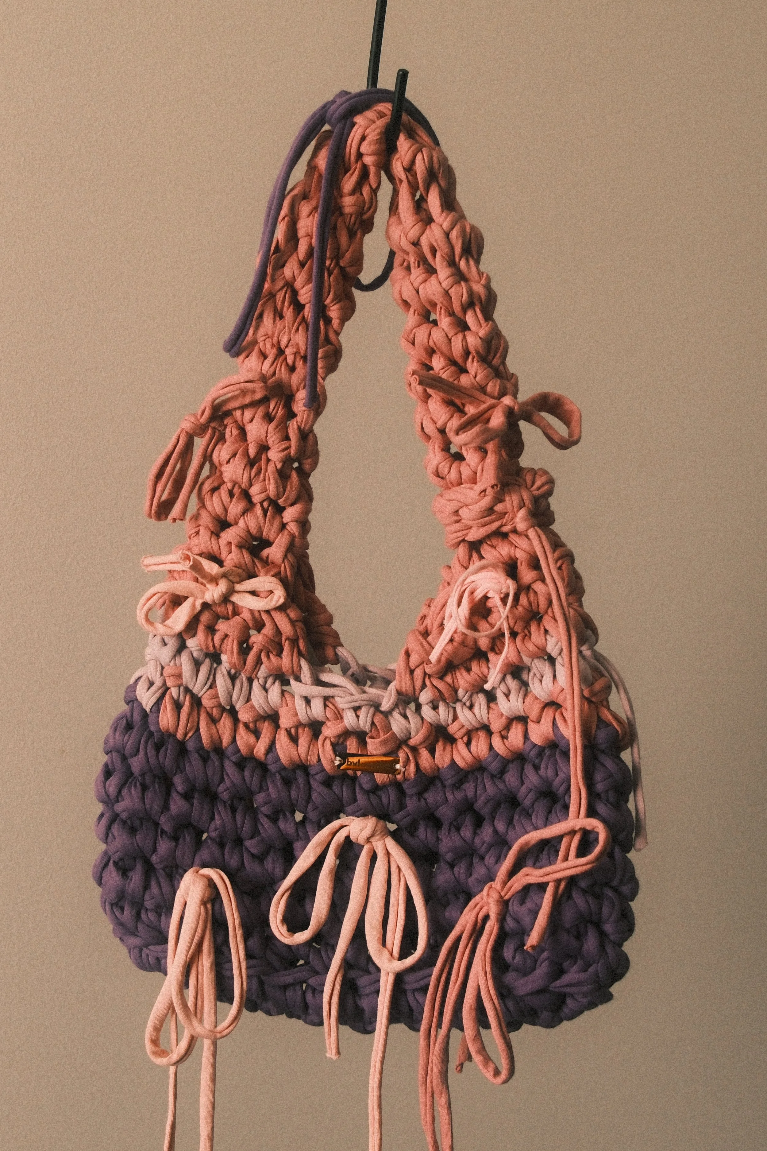 A handbag made of intertwined and braided yarn in shades of purple, pink, and beige, hanging from a hook against a plain beige background.
