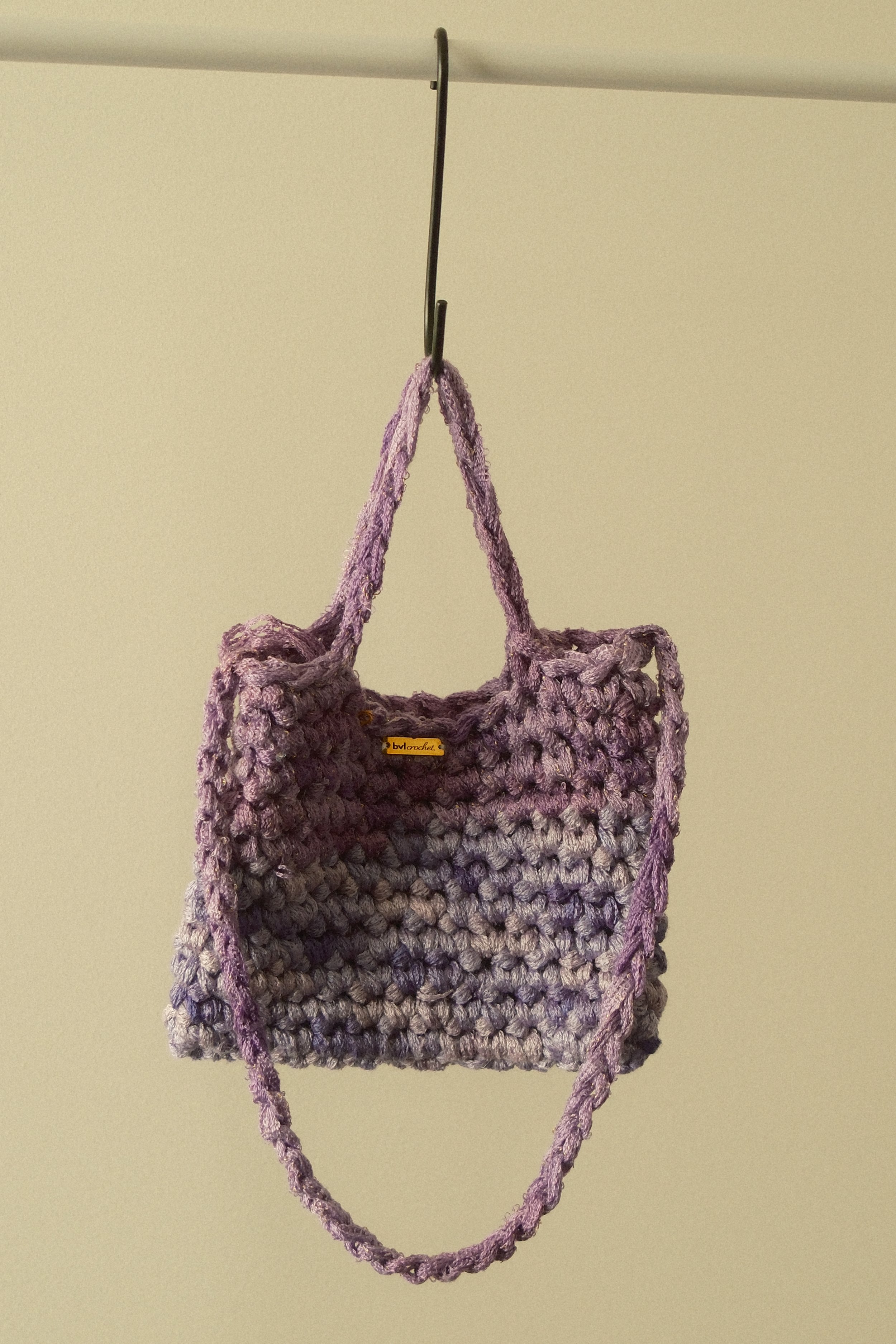 A handmade purple crochet bag hanging on a metal hook against a plain background.
