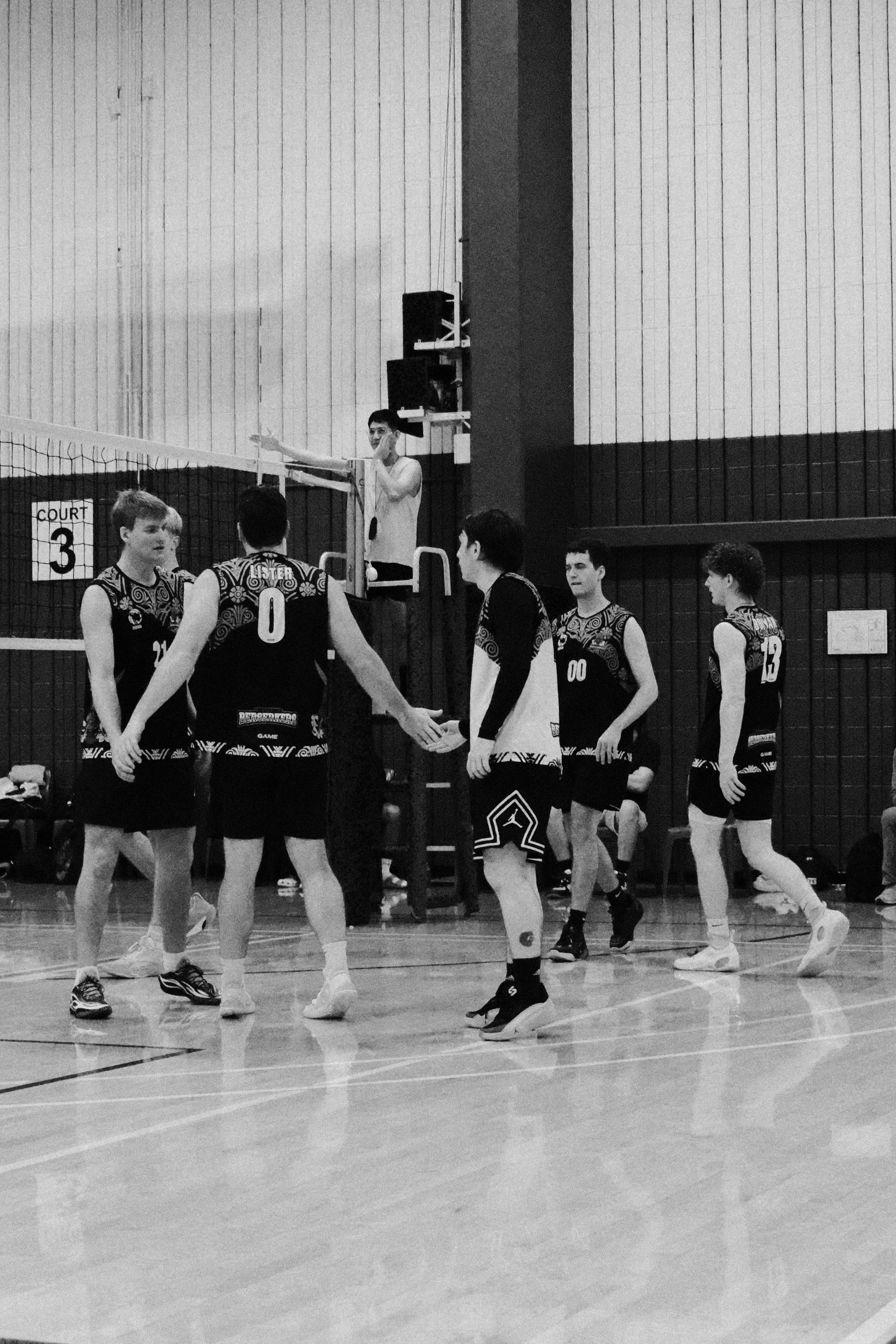A group of male volleyball players wearing matching jerseys holding hands in a gymnasium, likely during a game or practice, with a referee standing on a platform in the background.