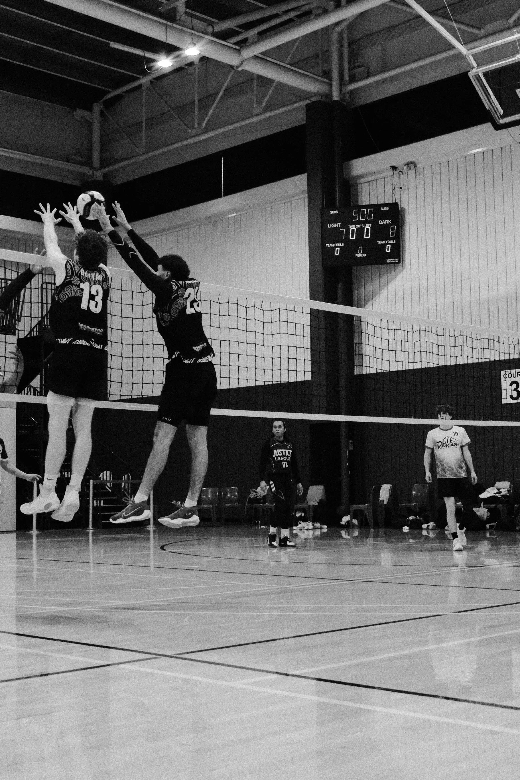 Two volleyball players, one wearing jersey number 13 and the other wearing number 20, jump to block a ball at the net during a game in an indoor gym. Several spectators or team members are visible on the sidelines, and a scoreboard shows 50 seconds left on the clock with the score tied at 7-7.