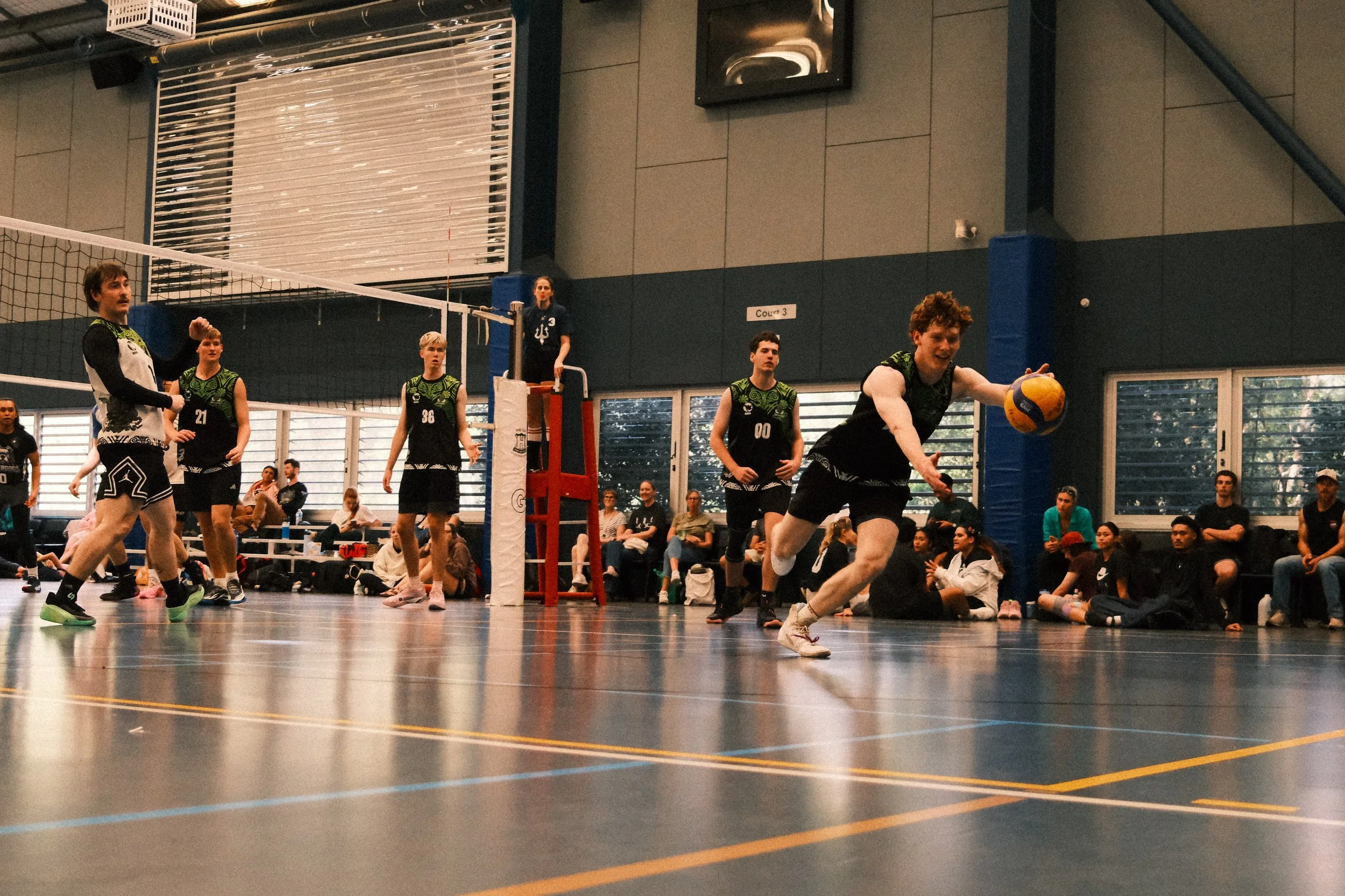 Indoor volleyball game with players on court, some preparing to serve or receive, spectators watching from the sidelines.