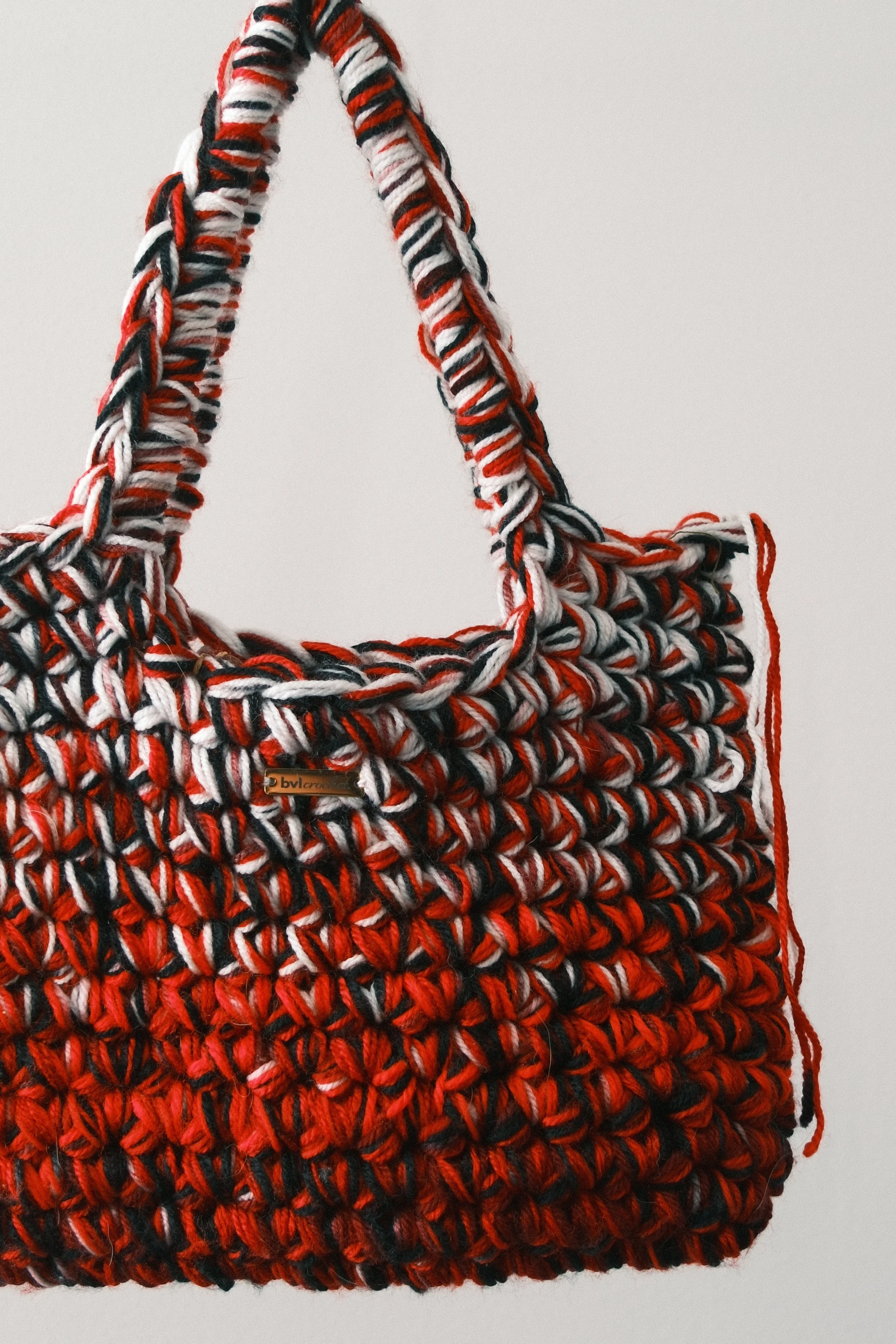 Close-up of a woven multicolored bag with red, black, white, and gray yarns, featuring a small leather tag with the brand name 'bvl cross'.