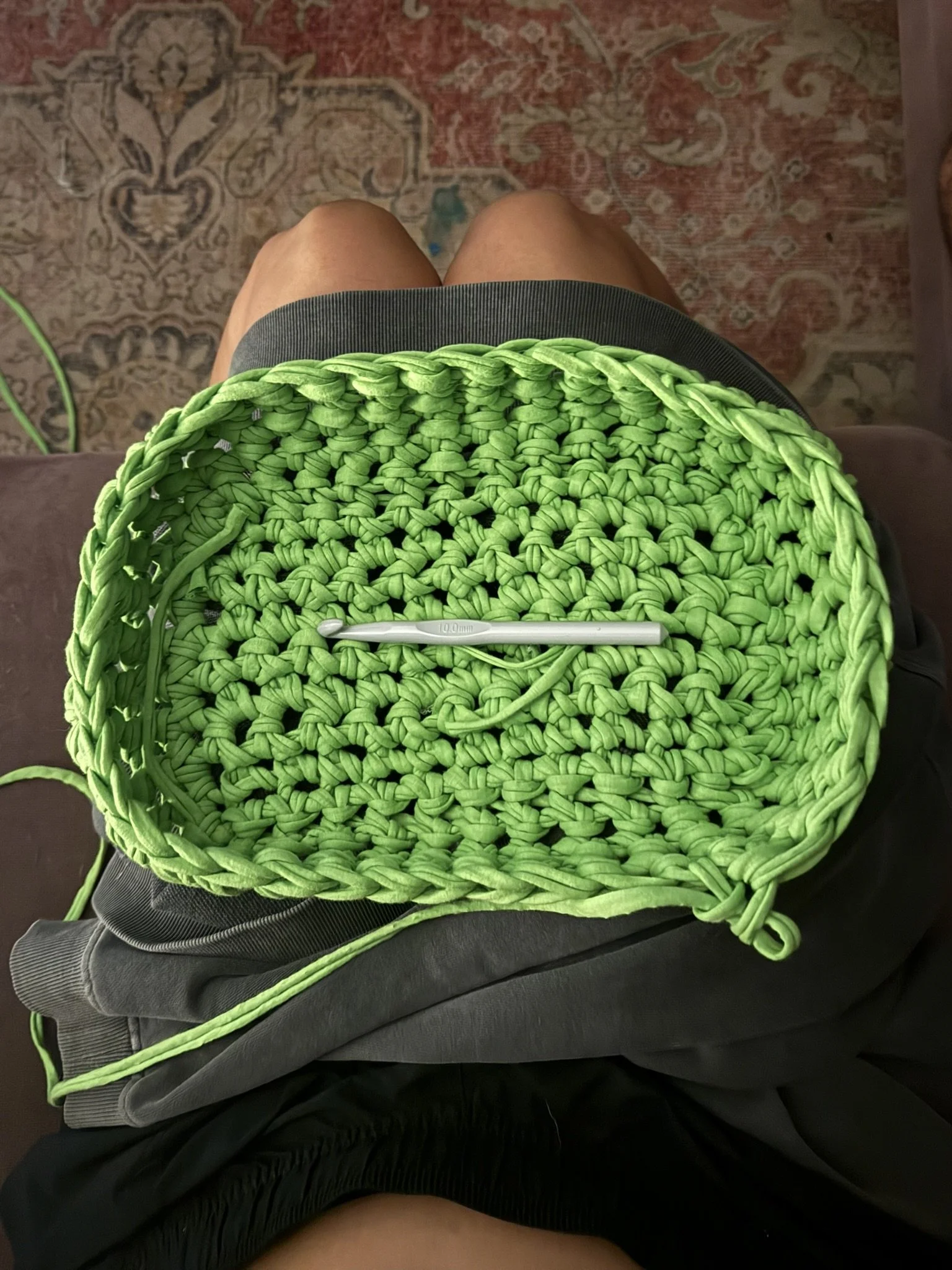 A person's lap with a crocheted green basket on top, a crochet hook resting inside it, and shorts and shorts covered with a black jacket.