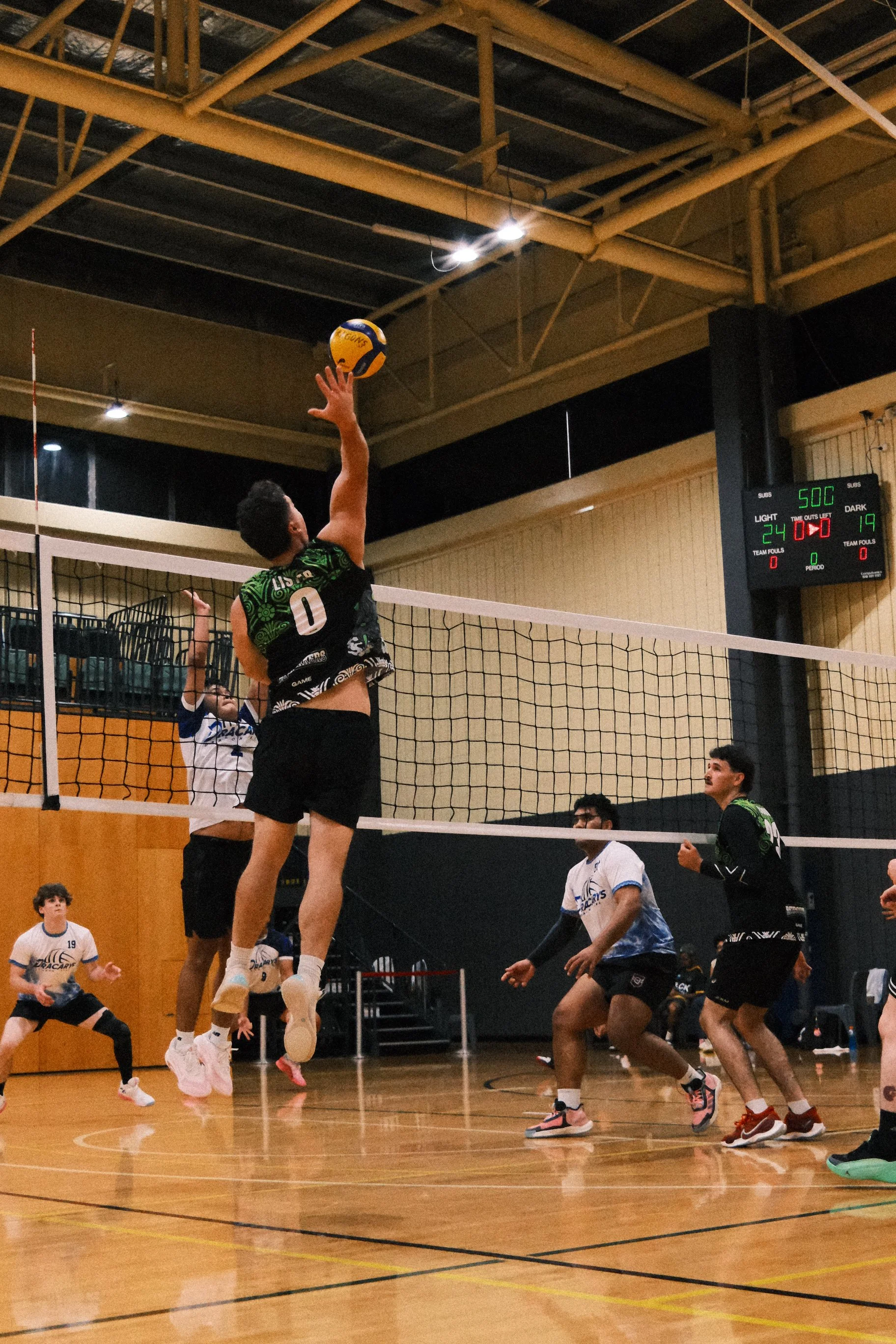 A volleyball game in progress with two players jumping to hit the ball at the net, surrounded by other players on an indoor court with a digital scoreboard showing scores and time remaining.