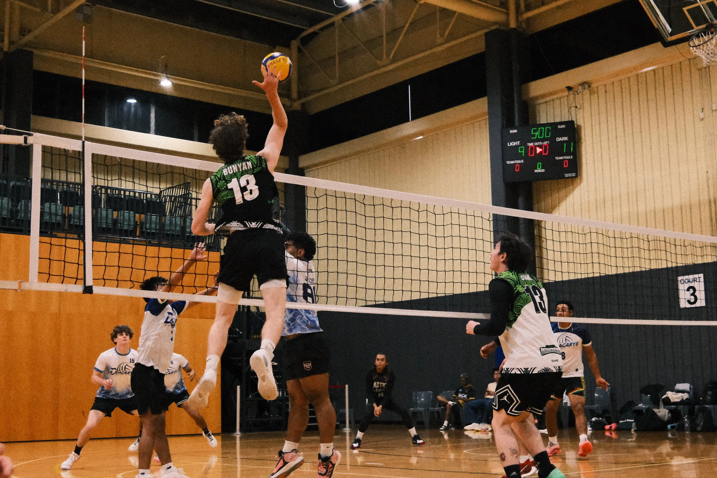 A volleyball game in progress with players jumping to hit the ball over the net. The player in the center wearing a black and green jersey with the number 13 is jumping to attack. Other players are positioned around the court, with some preparing to defend. The game is taking place indoors, with a scoreboard displaying a score of 9-11 and 5 minutes left on the game clock.