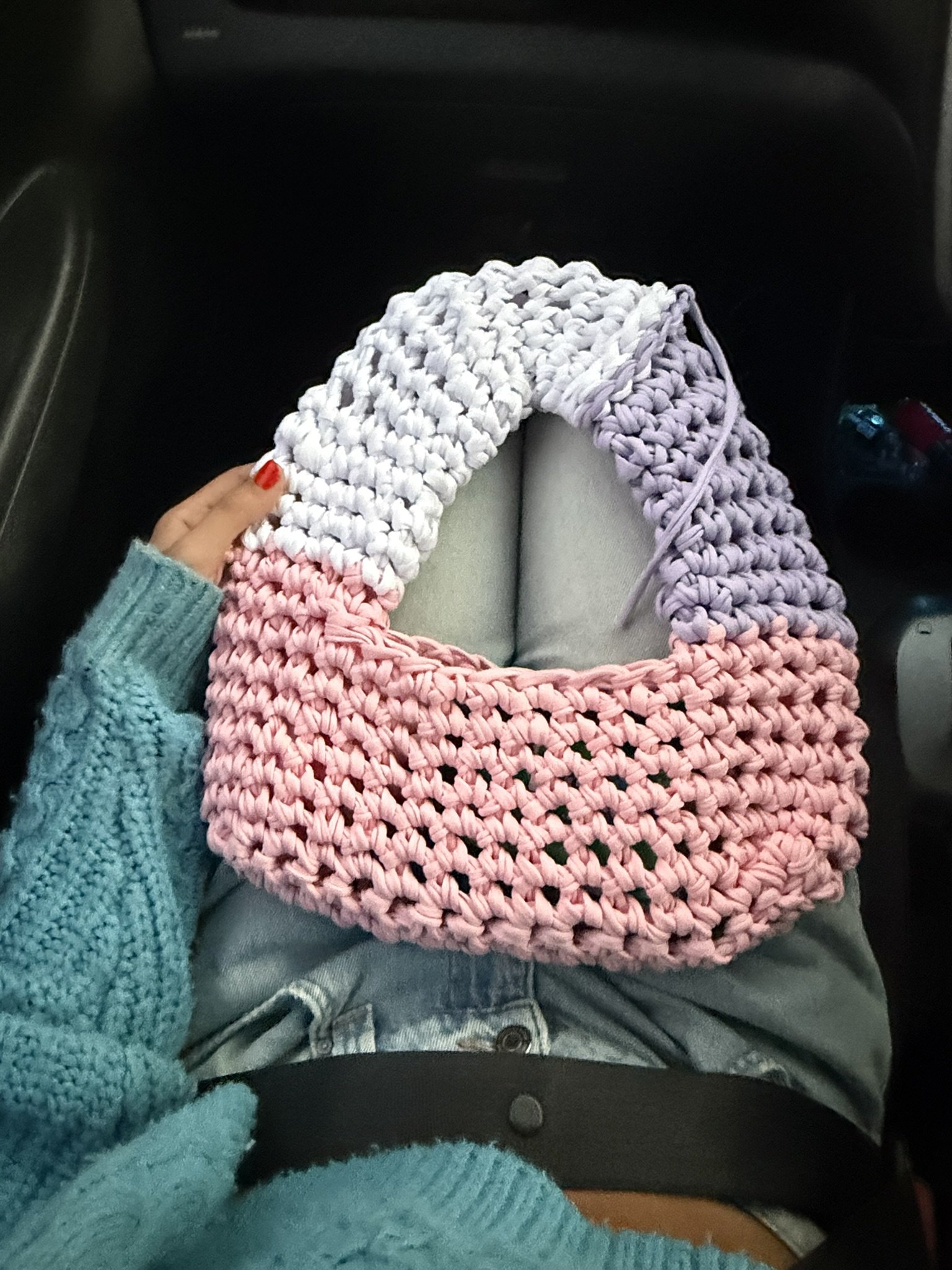 A person's lap with a pink, white, and purple crochet blanket, wearing light gray pants and a teal sweater, seated in a car.