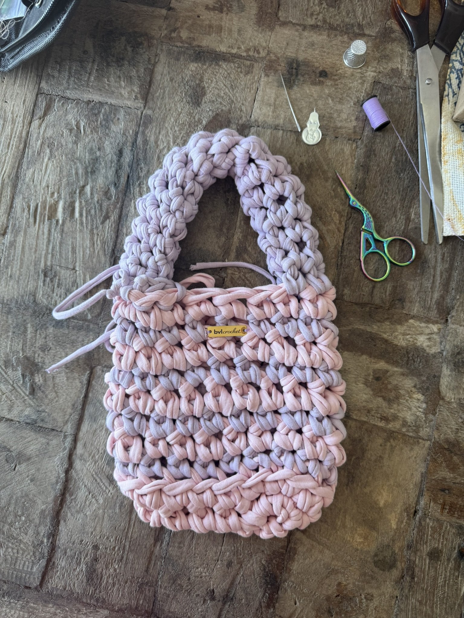 A pink chunky yarn crochet project resting on a wooden surface, with crochet tools and supplies nearby, including scissors, thread spools, a thimble, and a thimble-shaped pin or thimble case.