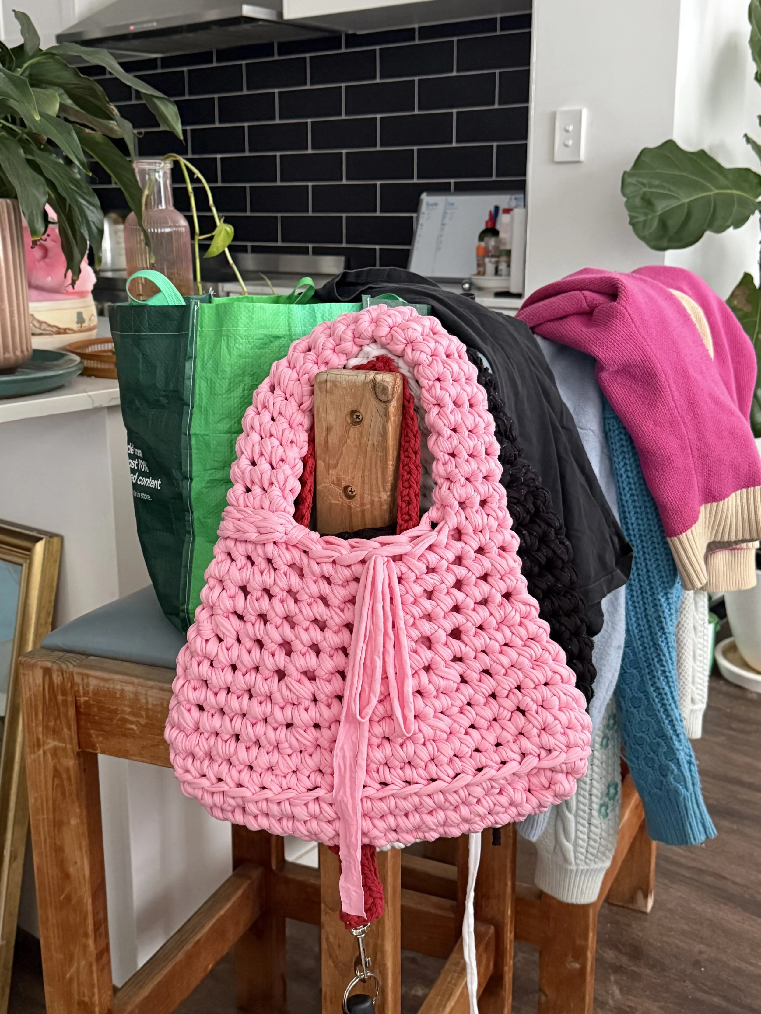 A pink crocheted handbag with a drawstring closure hanging from a wooden chair in a room with plants and various household items.