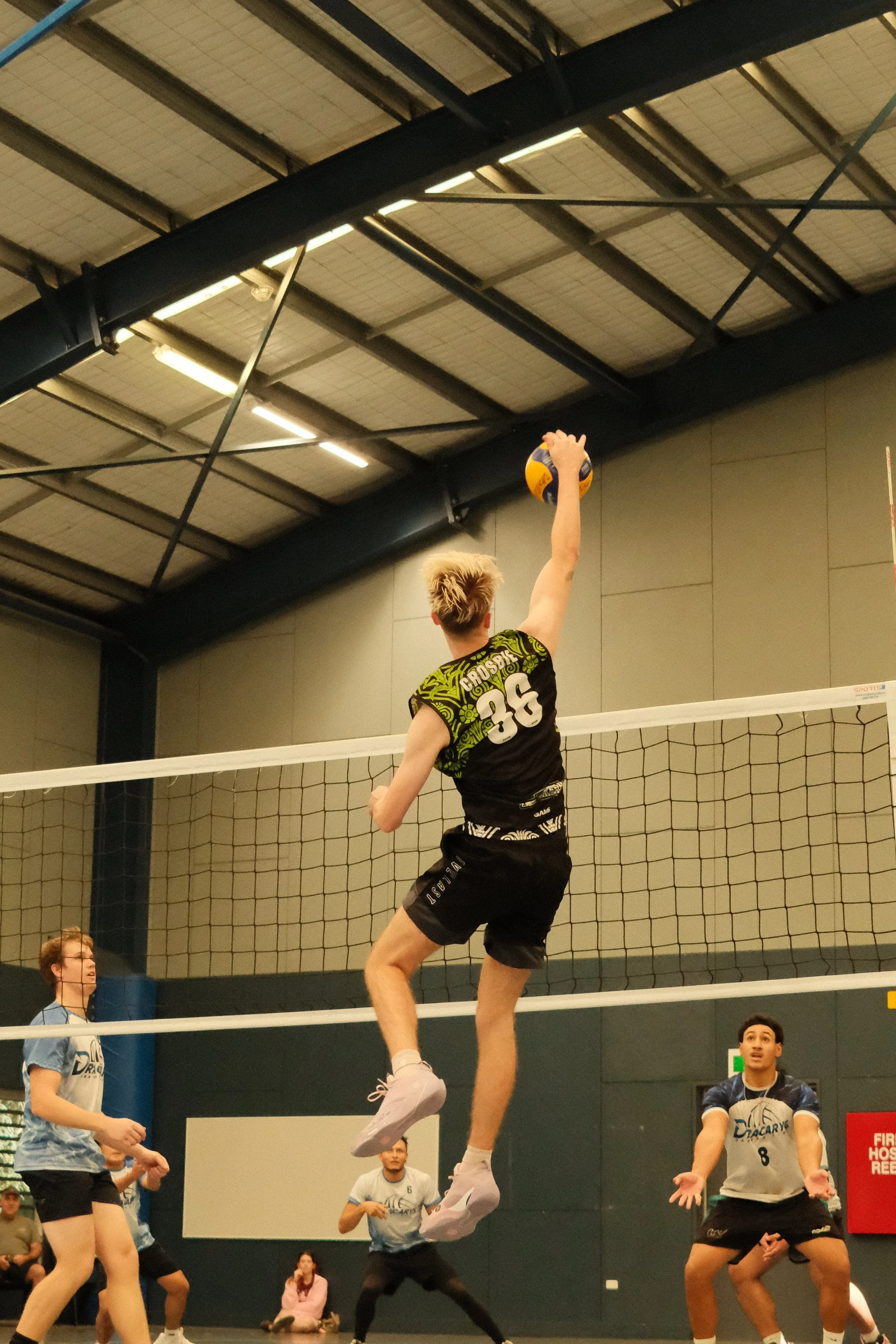 A volleyball player with light-colored hair jumps to hit the ball during a match, with other players and spectators watching in an indoor gym.
