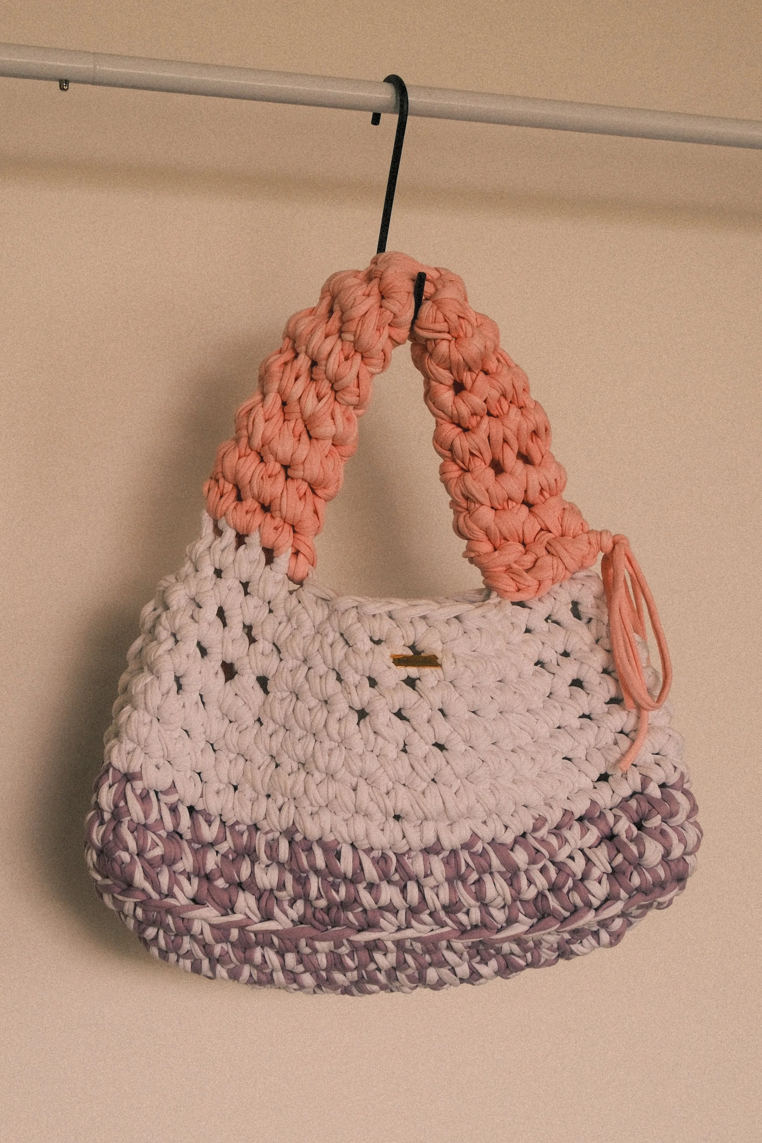 A handmade woven handbag with a rounded shape, featuring a gradient color pattern from white to pink and purple hues. The bag is hanging from a black hook attached to a metal rod against a beige wall.