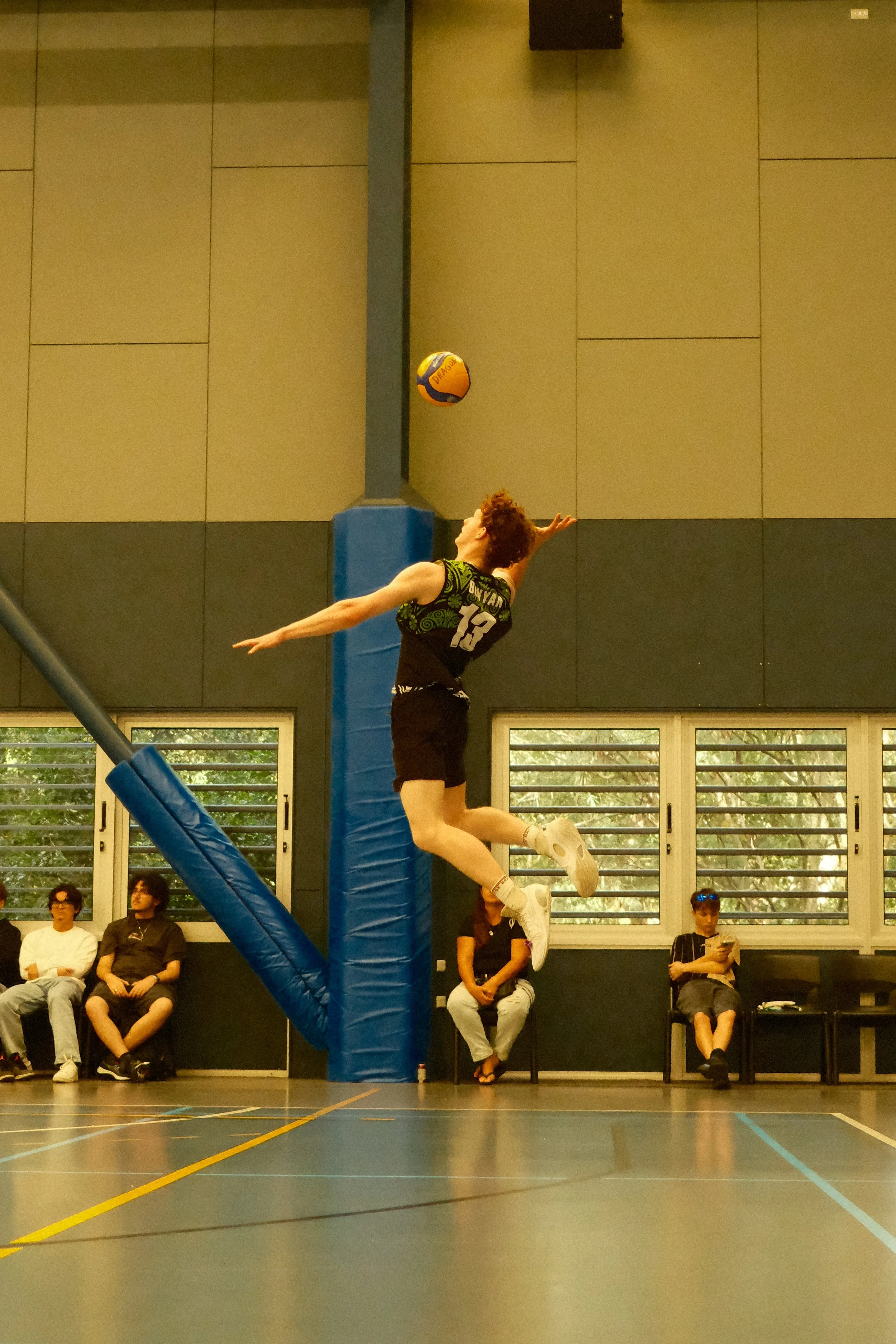 A female volleyball player with curly red hair, wearing a sports jersey and shorts, is airborne near the volleyball net, reaching for the ball during a game in an indoor gymnasium with spectators seated nearby.