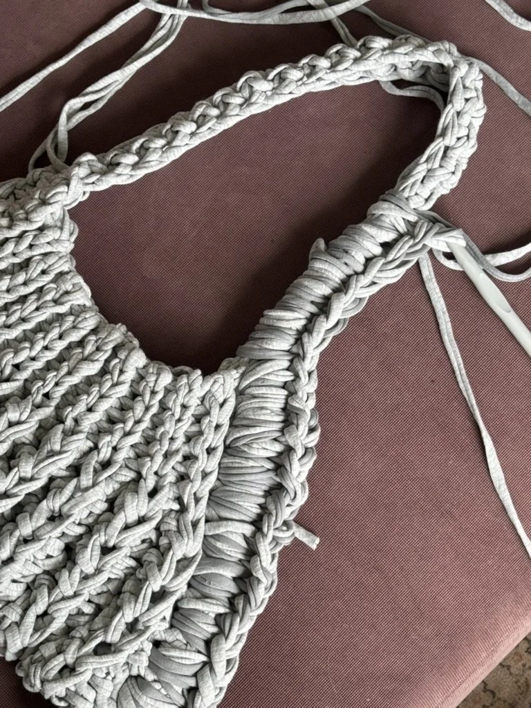 Close-up of a handmade woven bag made from light gray fabric strips, lying on a pinkish-red surface.