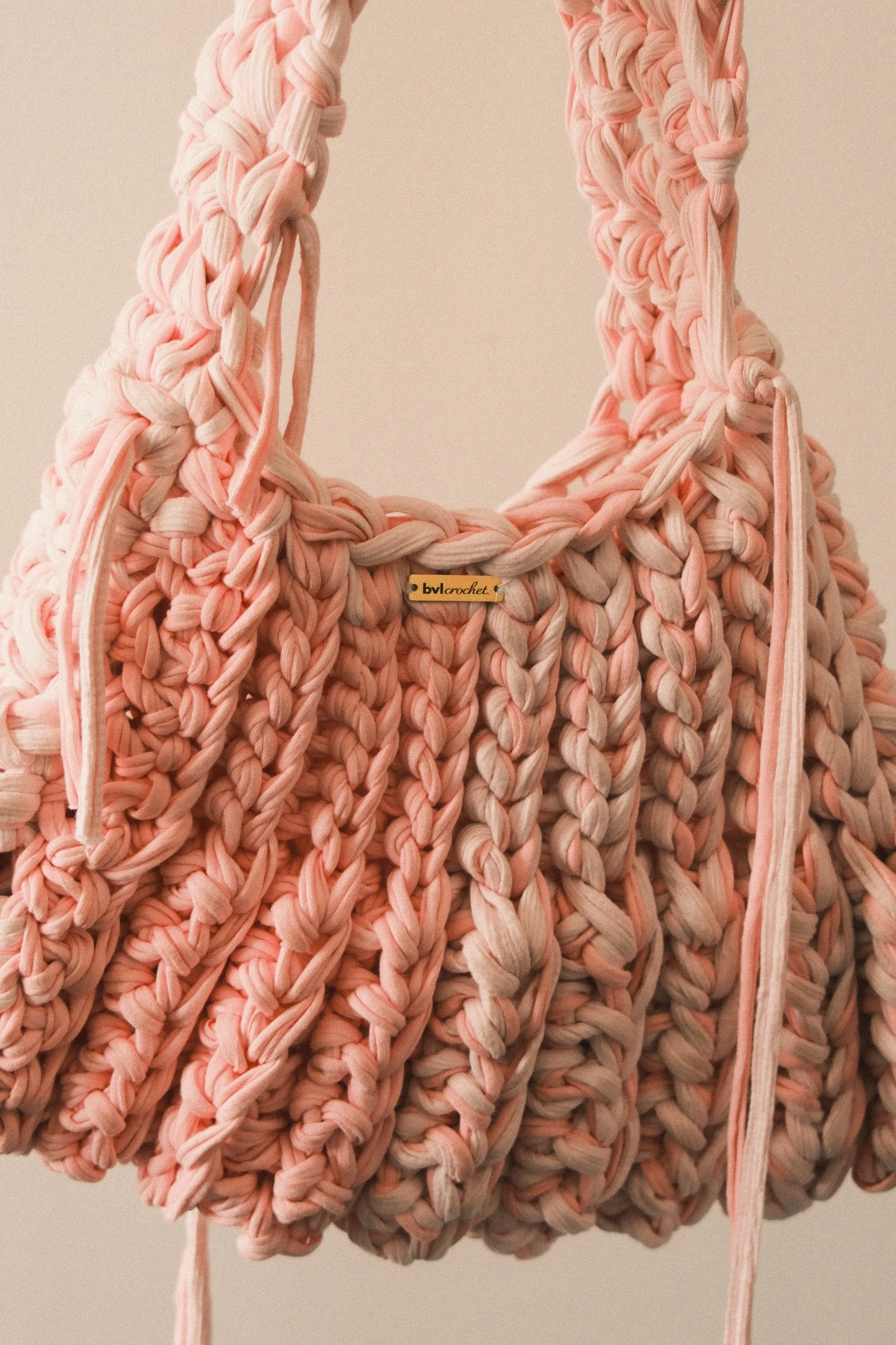A close-up of a large, pink, hand-crocheted bag made from thick yarn, with a braided handle and a small tag that reads 'bvl crochet'.