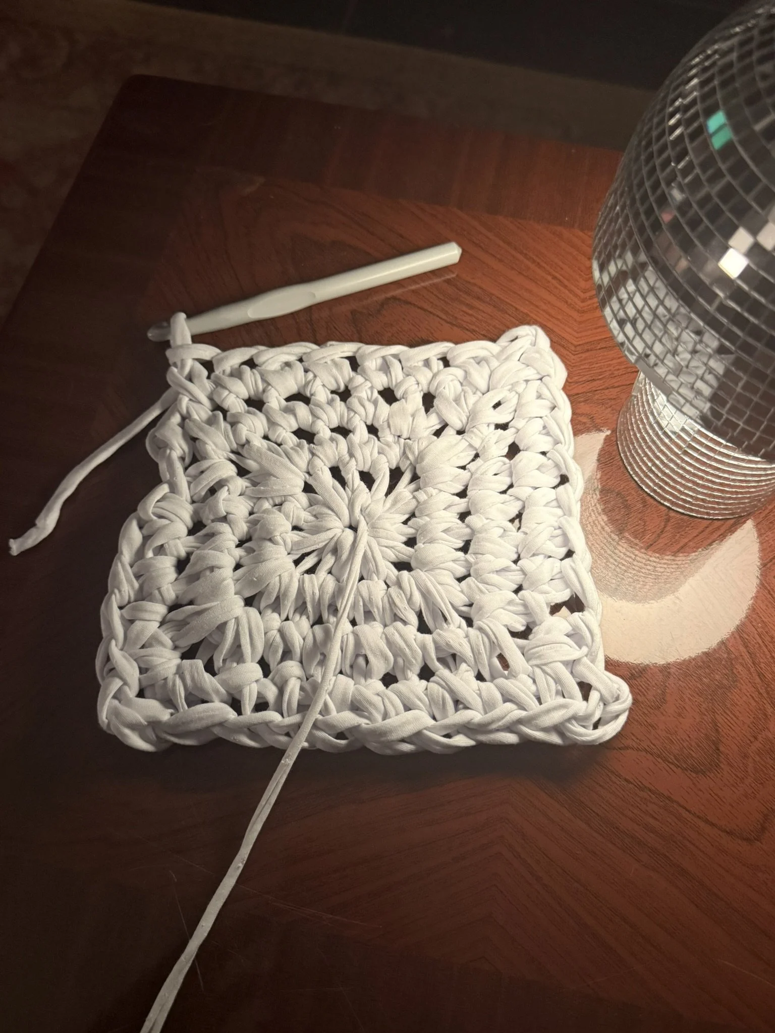 Close-up of a wooden table with a white crochet square, a white plastic tool, a silver disco ball, and a small portion of a wooden chair.