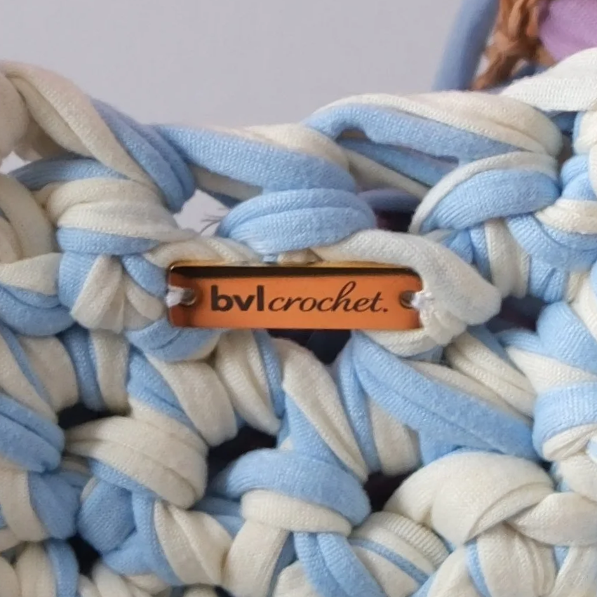 Close-up of a crochet project made with blue and white fabric strips, with a small orange tag reading 'bvl crochet' attached to the fabric.