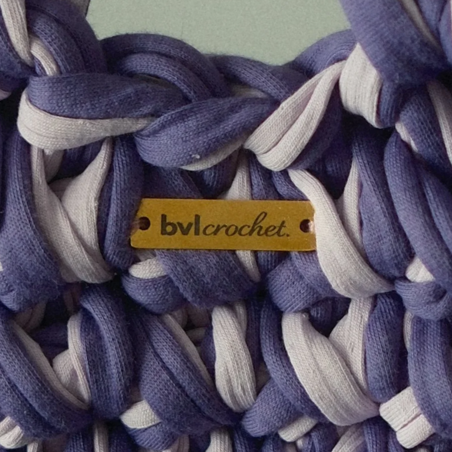 Close-up of a woven scarf or fabric with purple and white stripes, attached to a small yellow tag that reads 'bvlcrochet'.