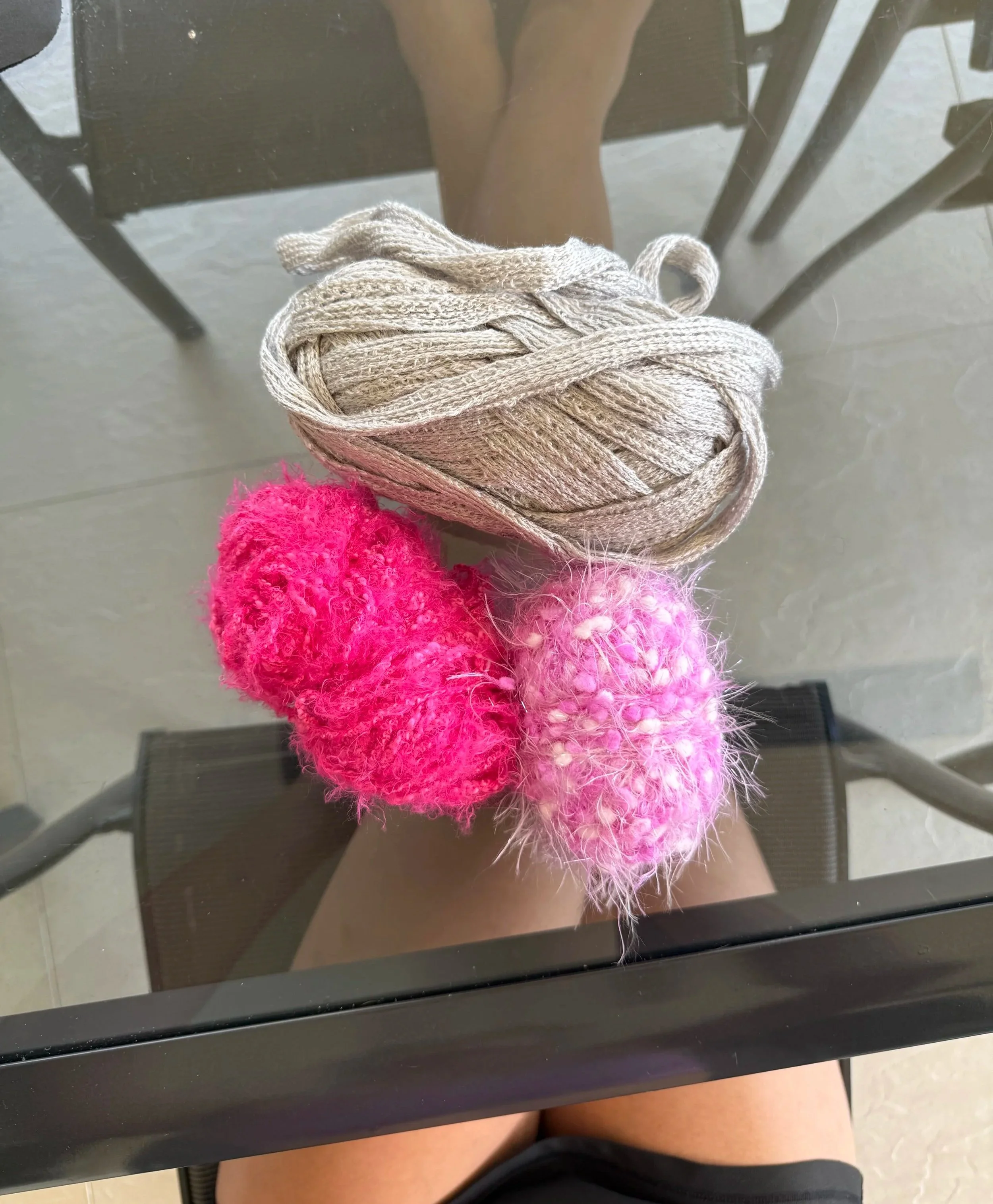 Reflective glass table showing a coil of beige yarn, two fuzzy pink pom-pom decorations, and part of a person's legs.