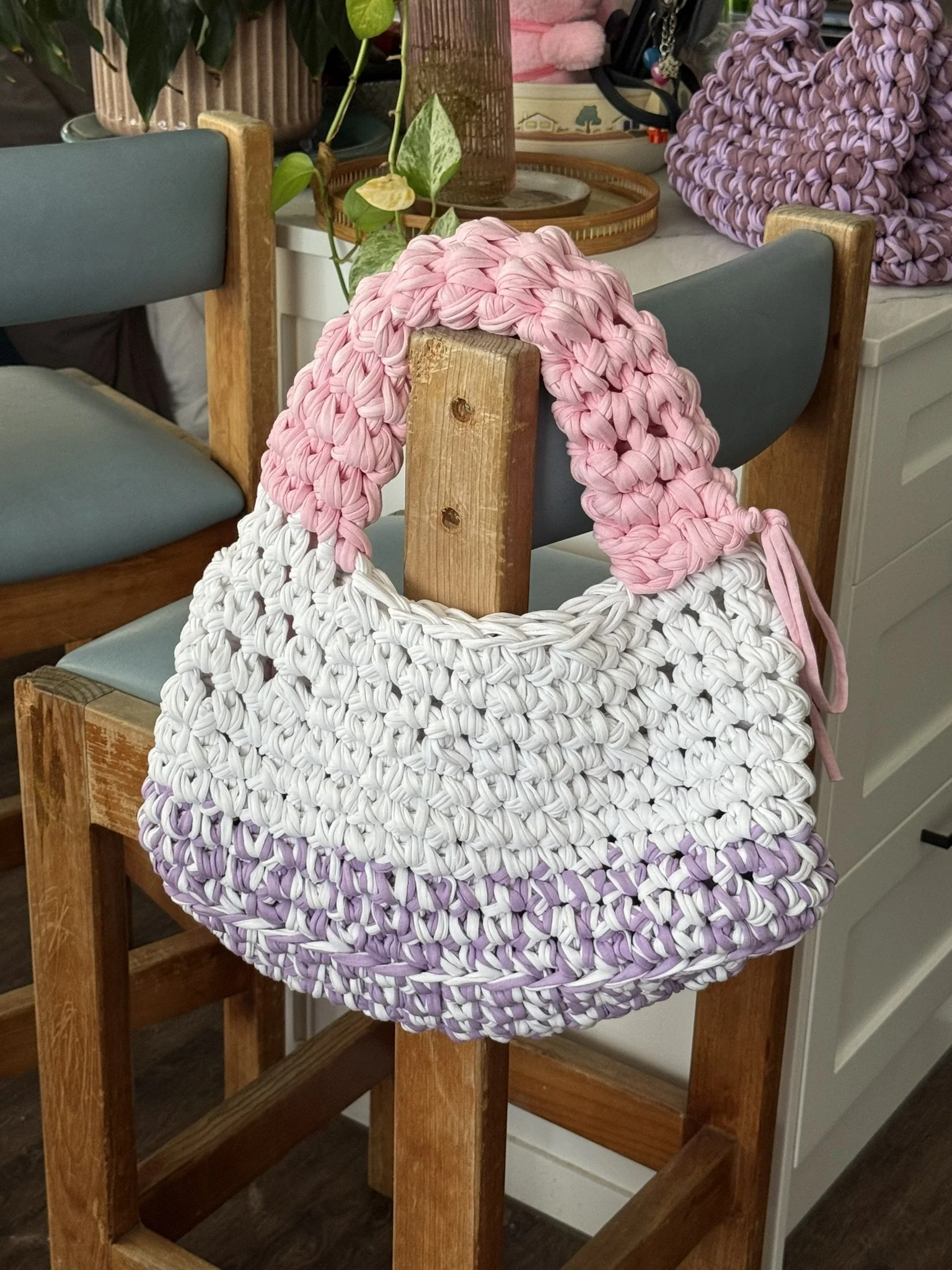 A woven bag made of intertwined pink and white fabric hangs on the back of a wooden dining chair in a cozy kitchen or dining area. The chair has gray cushions and the area is decorated with potted plants and other kitchen items.