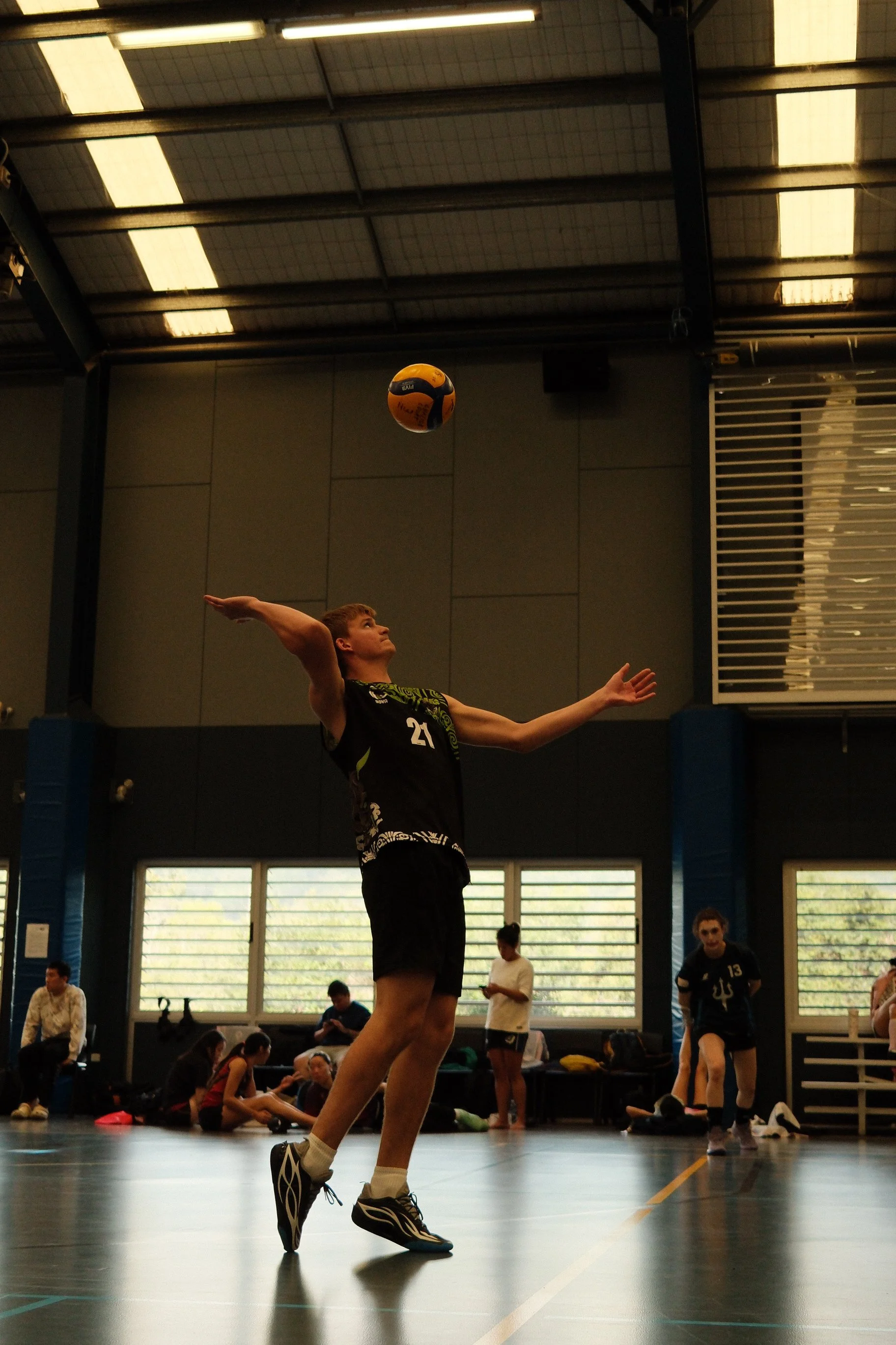 A young male volleyball player in a black jersey and shorts is jumping and preparing to serve the ball during a game or practice in an indoor gymnasium. Other players and spectators are visible in the background.