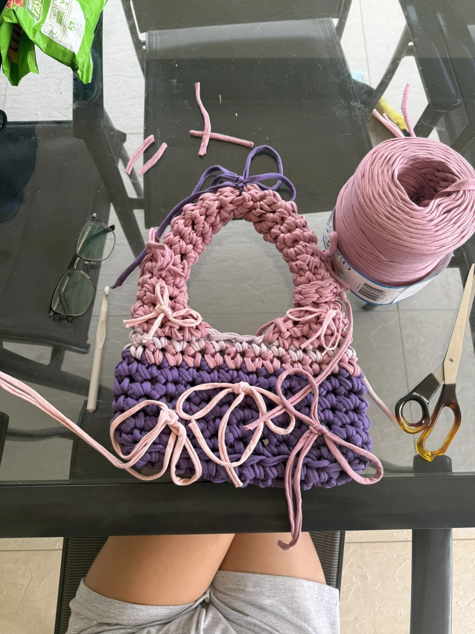 A table with knitting yarn, knitting tools, and glasses on it. The person is sitting with legs crossed, wearing gray shorts, and is working on a rainbow-colored knitted project.