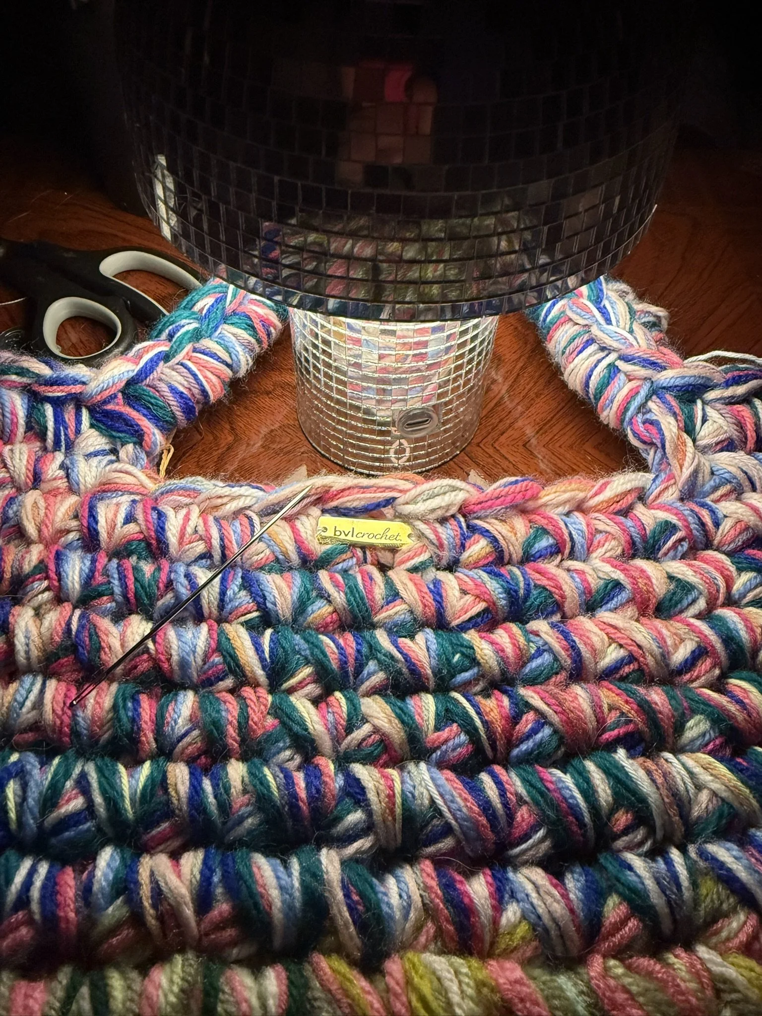 Close-up of colorful yarn strands surrounding a small crochet hook, with a mirrored disco ball in the center and a large black mirror in the background.
