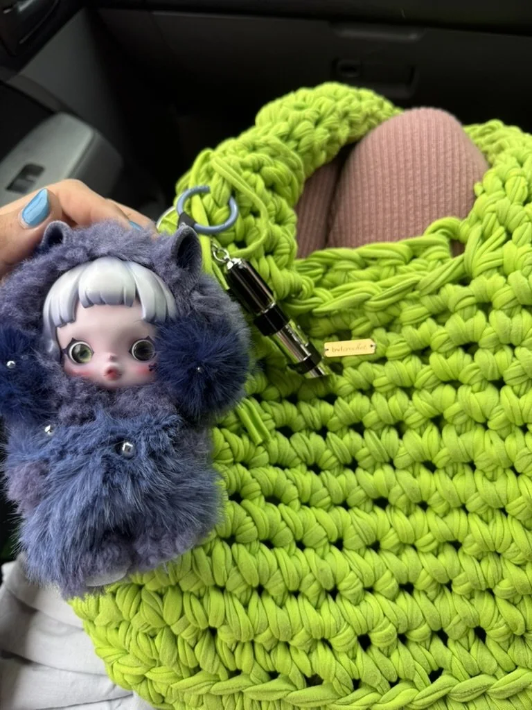 A hand holding a doll with a large head, big eyes, and silver hair, attached to a furry blue keychain or bag charm. The background shows a person sitting with pink pants in a bright green, woven bag or blanket, in a vehicle.