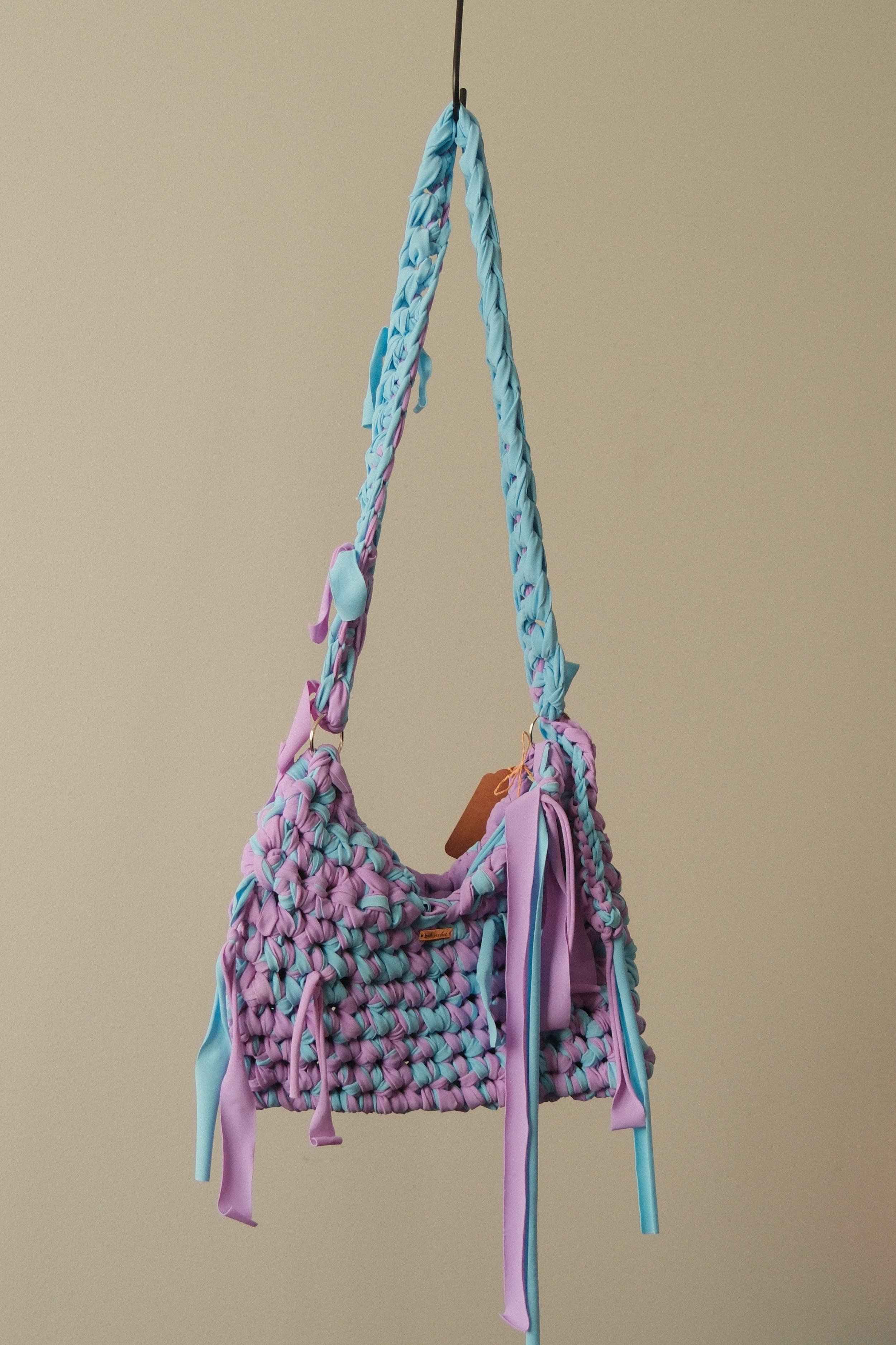 Colorful woven handbag made of pink, purple, and turquoise fabric with dangling fabric straps and a small brown tag, hanging from a black hook against a beige background.