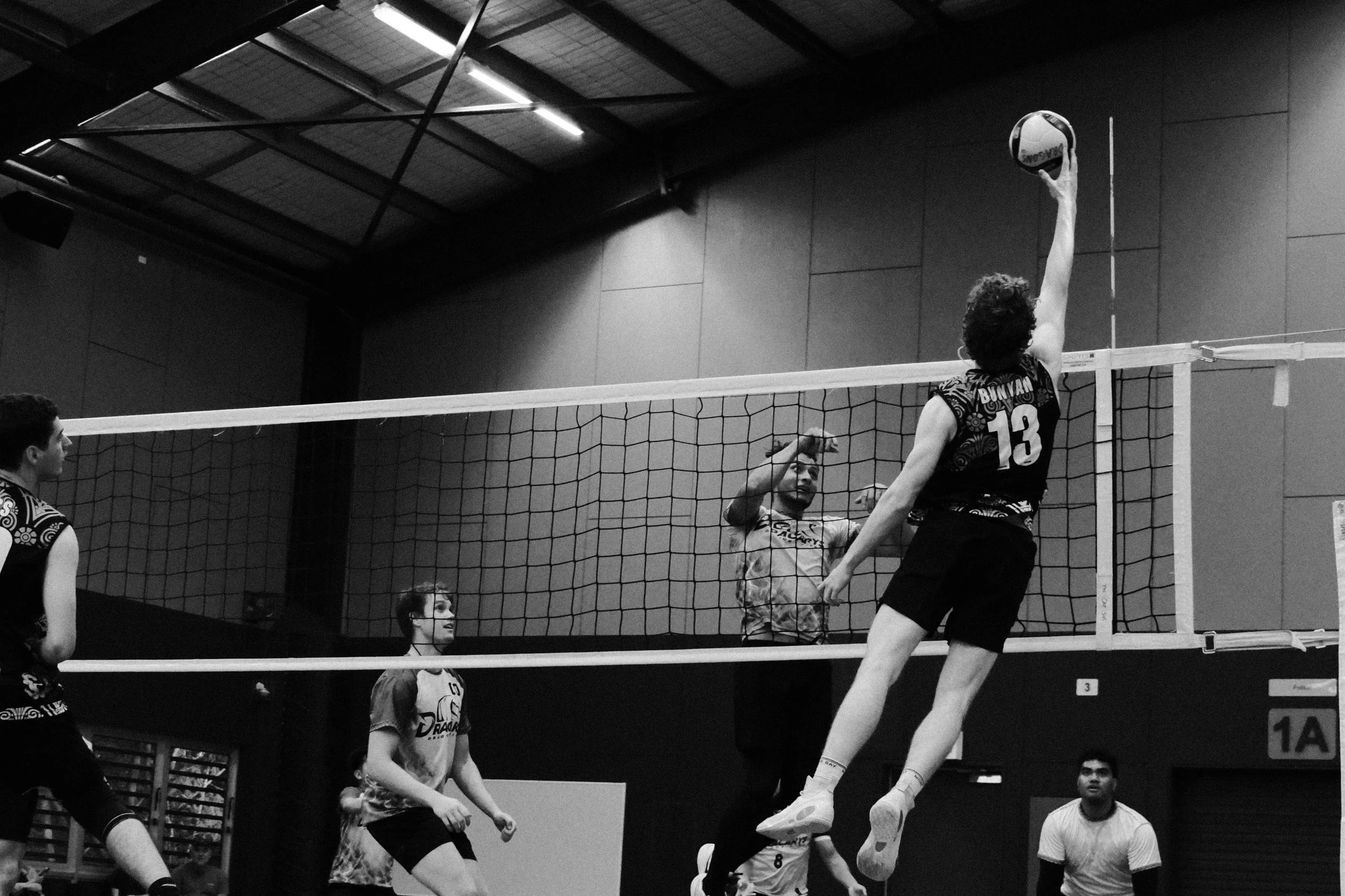 A black-and-white photo of a volleyball game in progress inside a gymnasium. A player wearing jersey number 13 is jumping to spike the ball over the net, while a player on the opposing team attempts to block it. Several spectators are watching, and a referee is visible in the background.