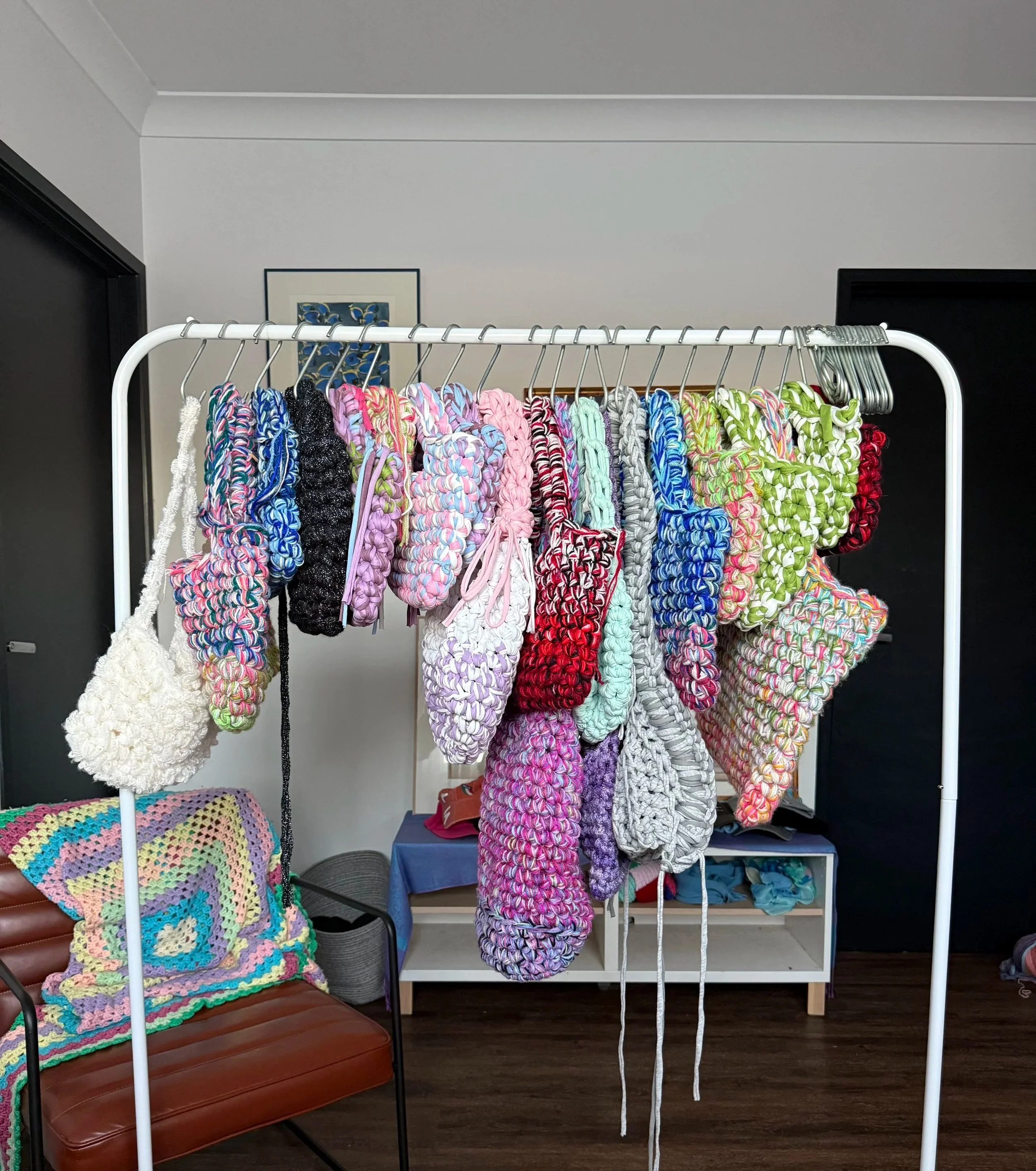 A display of colorful handmade crochet bagels hanging on a white clothing rack, with a knitted blanket on a chair and various bags and clothing in the background.