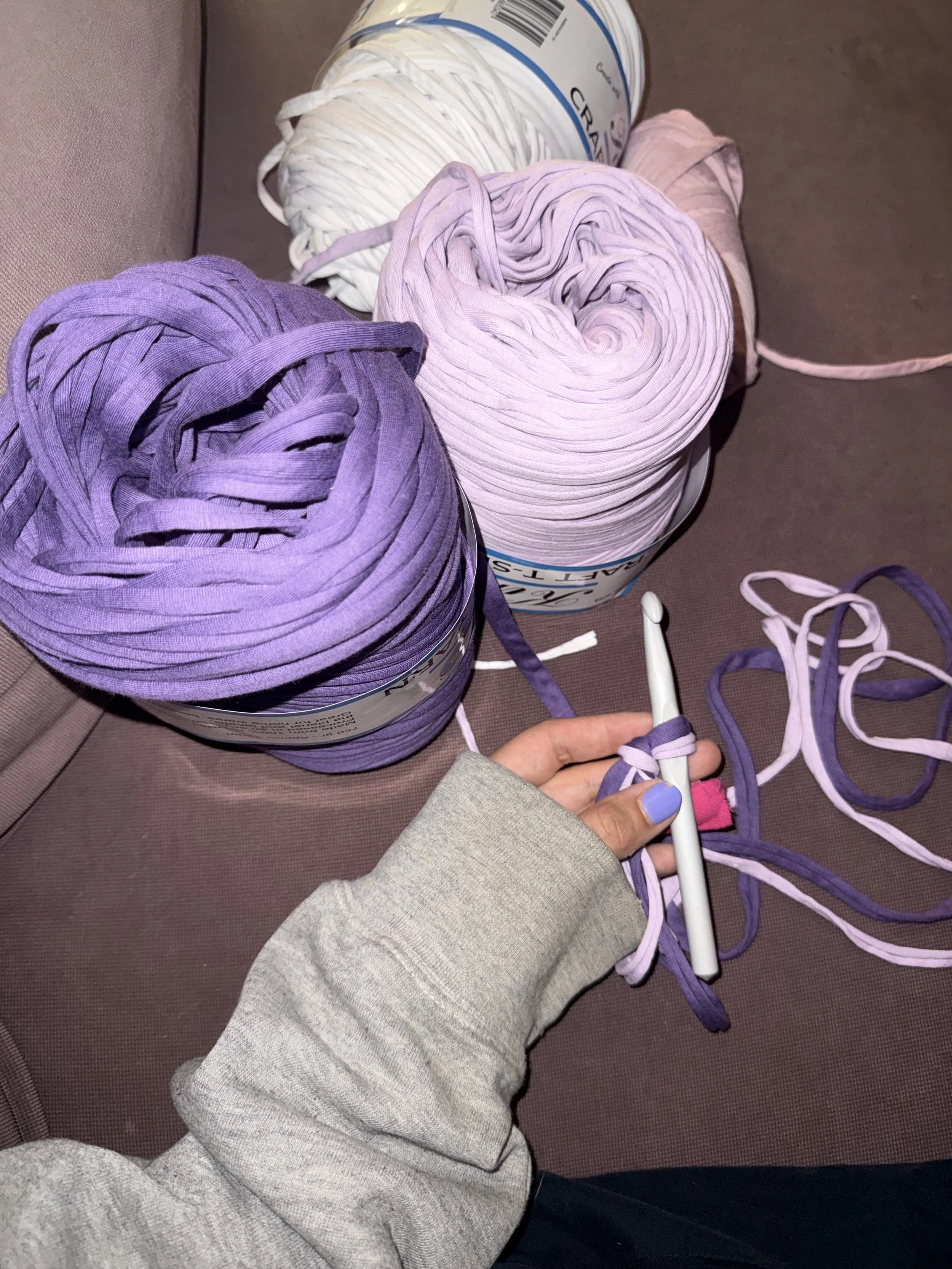 Close-up of purple and pink yarn balls on a couch, with a person's hand holding a knitting needle and knitting the purple yarn.