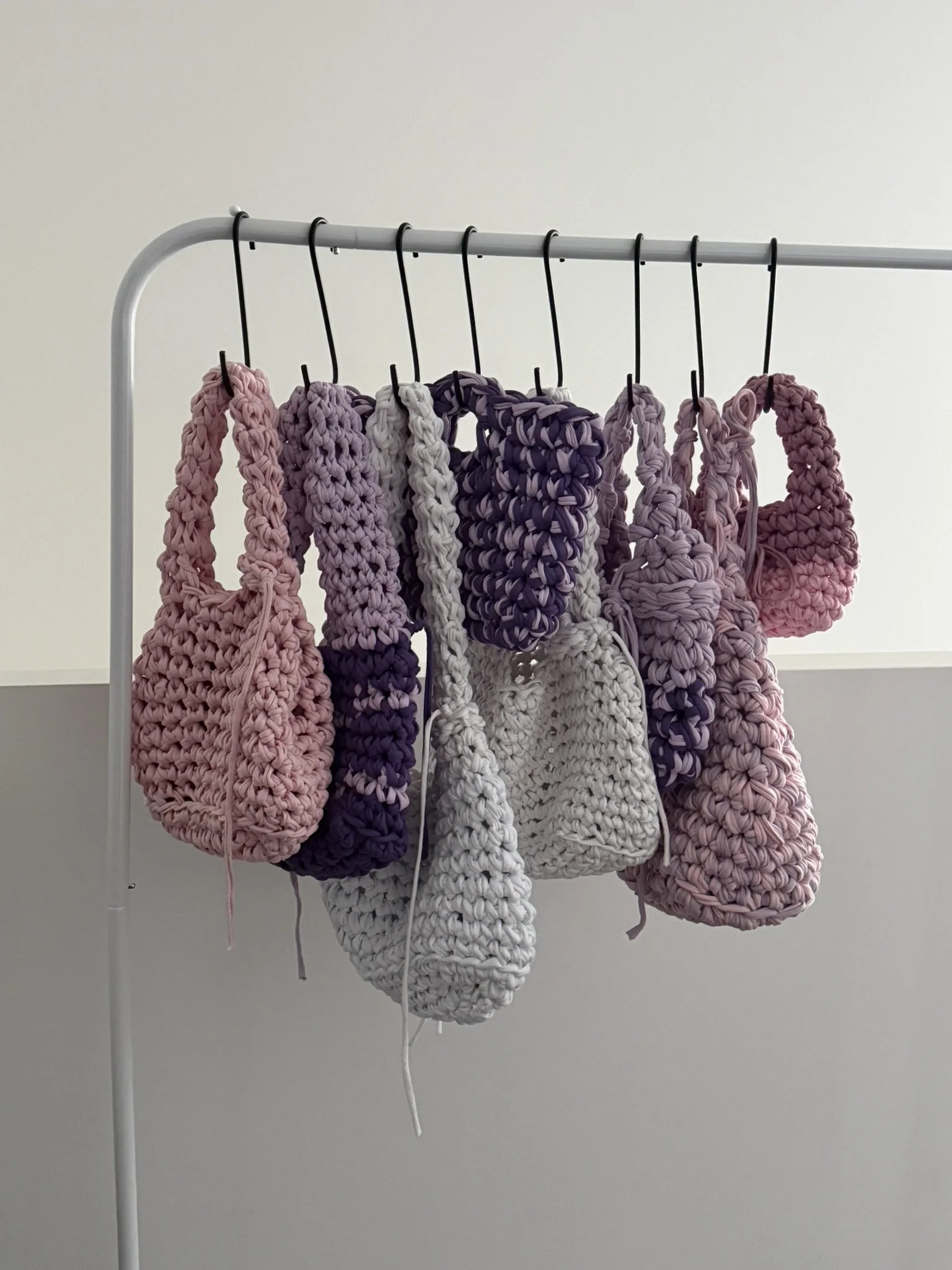 Multiple hand-knit scrunchies in shades of pink, purple, and white hanging on black hooks from a white metal clothing rack against a plain gray wall.